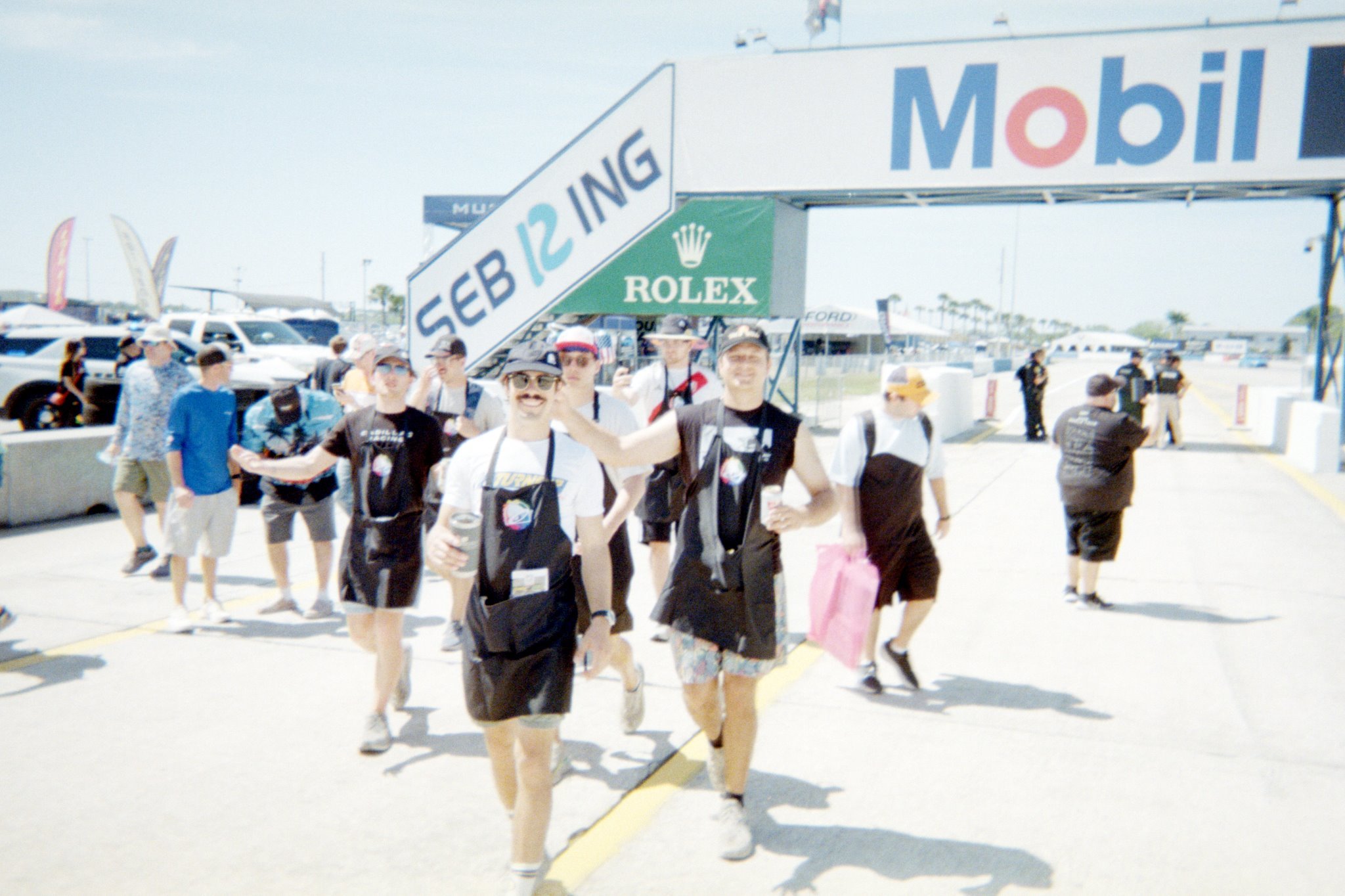People walking at a racing event under a large sign that says 'SEB 12 ING' with logos for Rolex and Mobil.