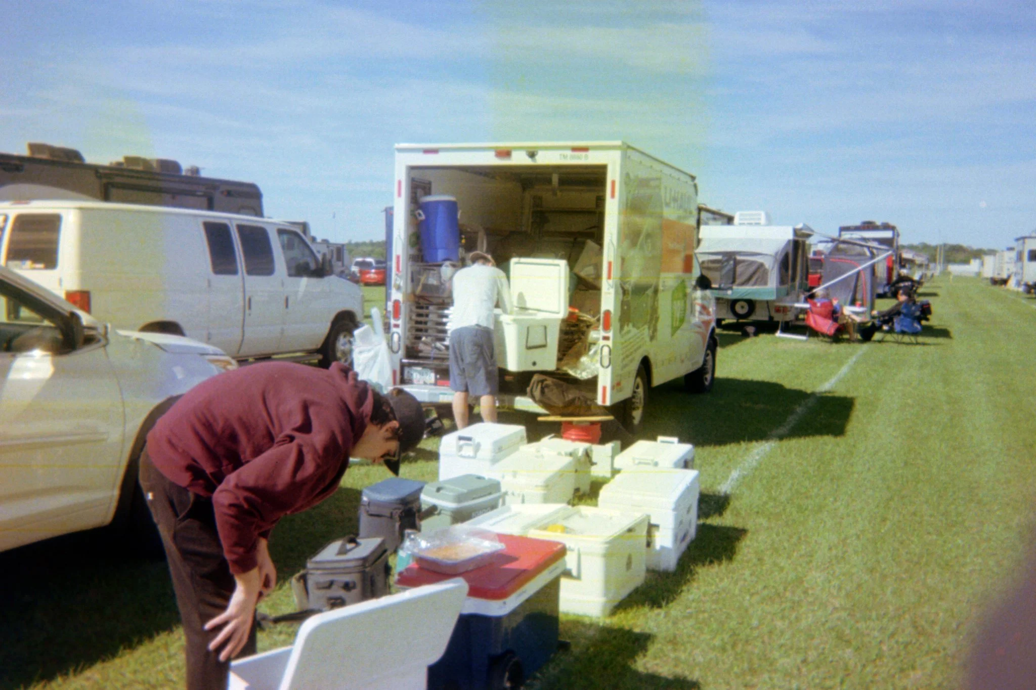 People setting up food and equipment at an outdoor event near parked vehicles on a grassy field.
