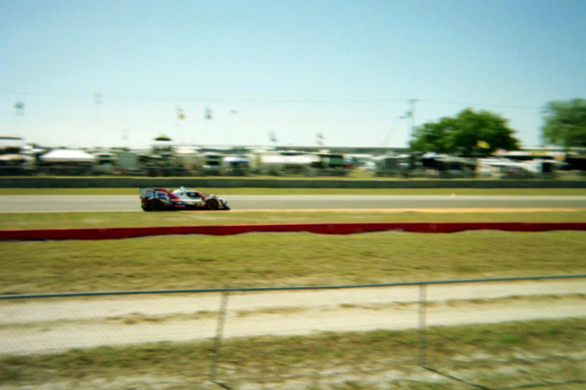Speeding race car on a track during a sunny day with a blurred background of tents and trees.