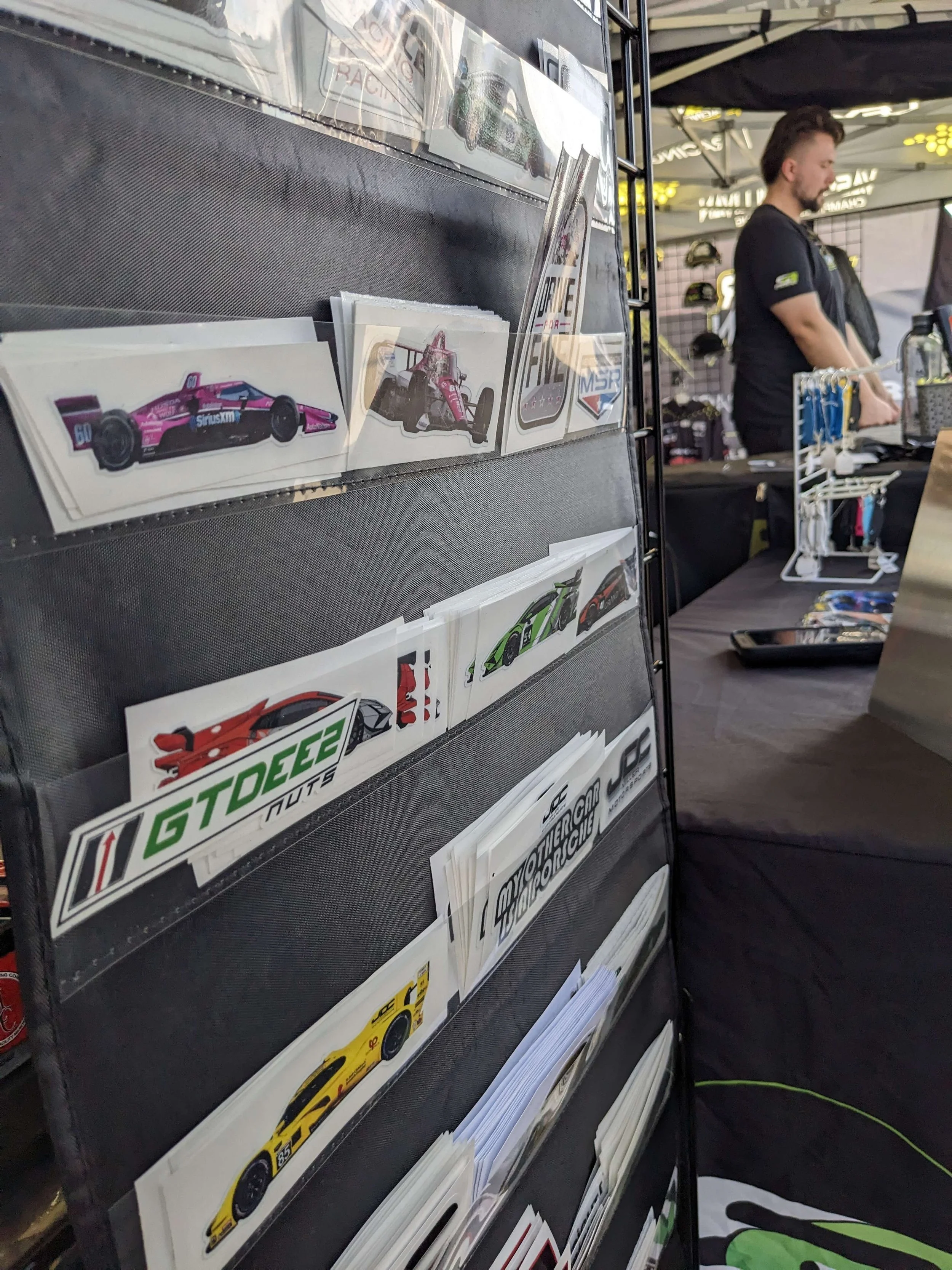 A display rack filled with stickers featuring various race cars, set up at a booth with a person standing behind a table.