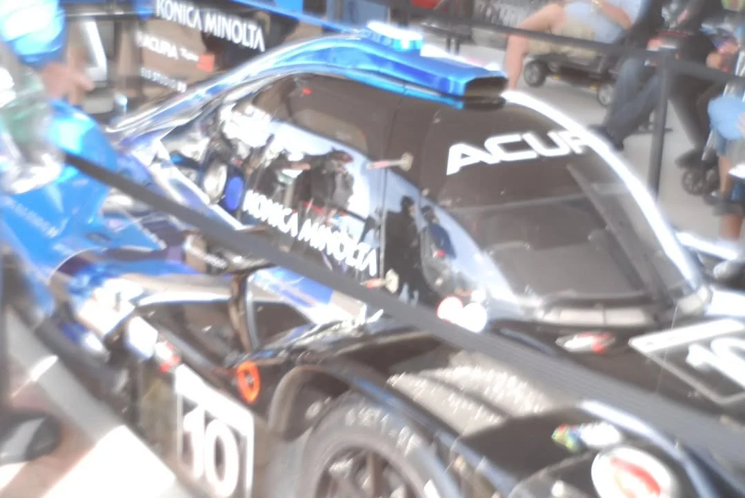 Blurred image of a blue and black race car with various sponsor logos, including Acura and Konica Minolta, surrounded by people at what appears to be a racing event or exhibition.
