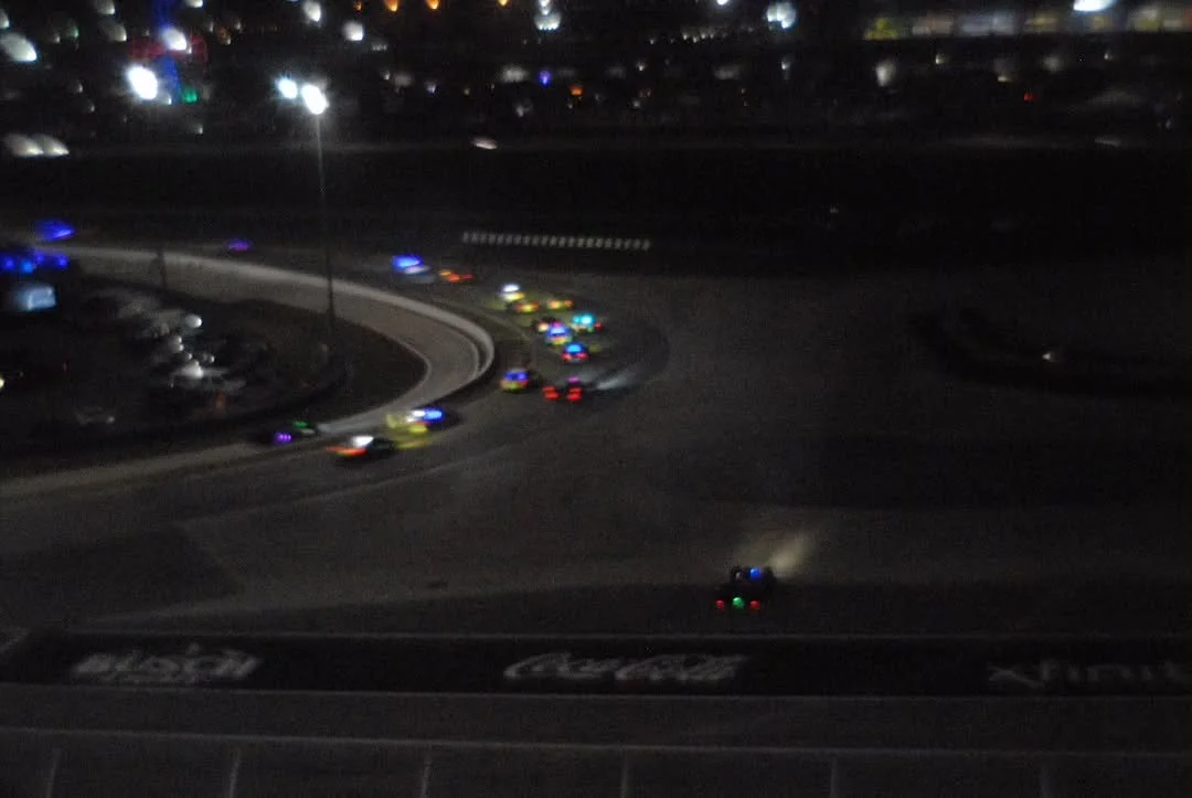 Nighttime scene viewed from a vehicle with a dashboard, showing a curved road with several police cars with flashing lights and a line of cars in a parking lot.
