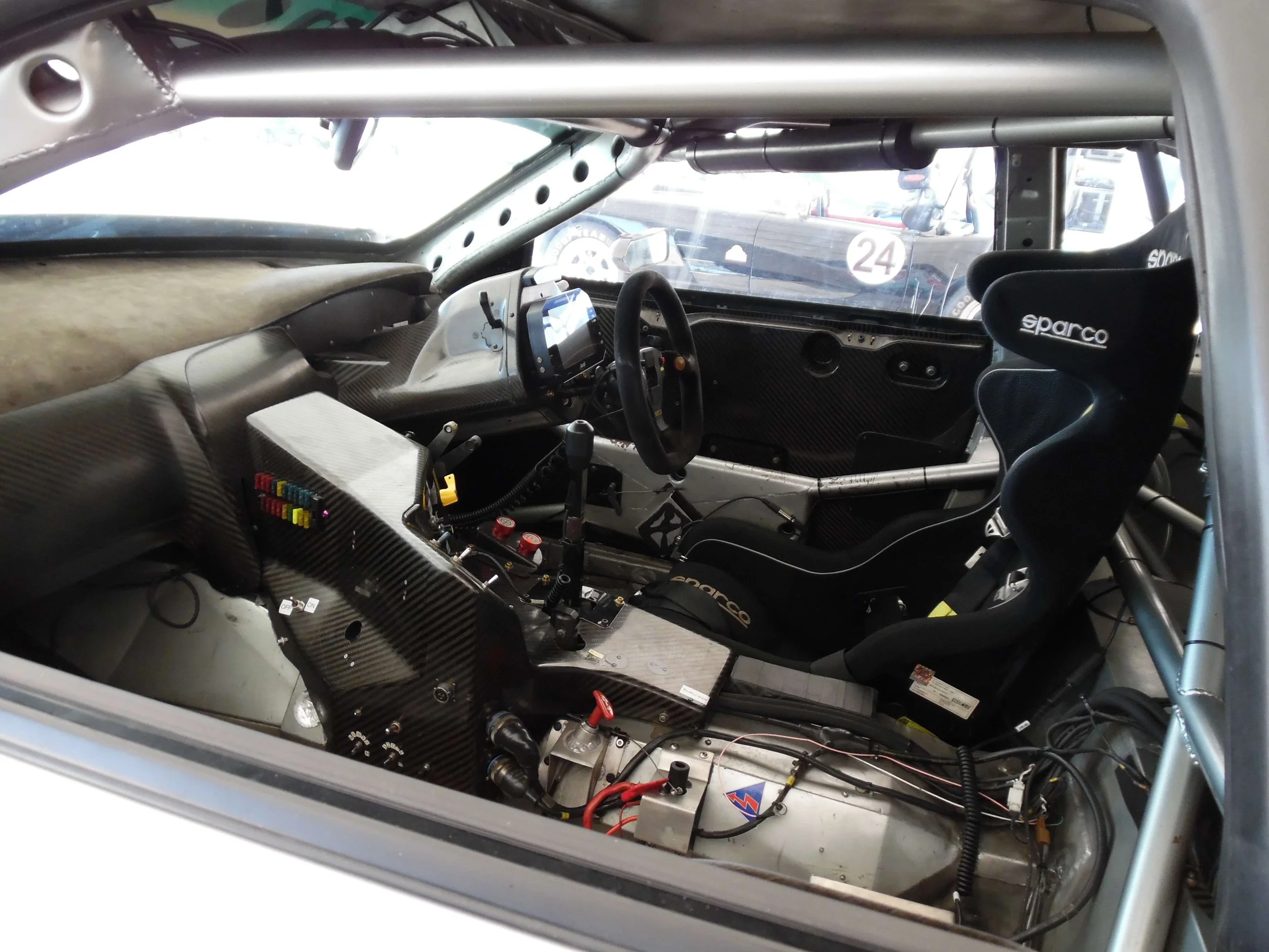 Interior of a race car featuring a steering wheel, racing seat with harness, dashboard with digital display, and various control buttons and wires.
