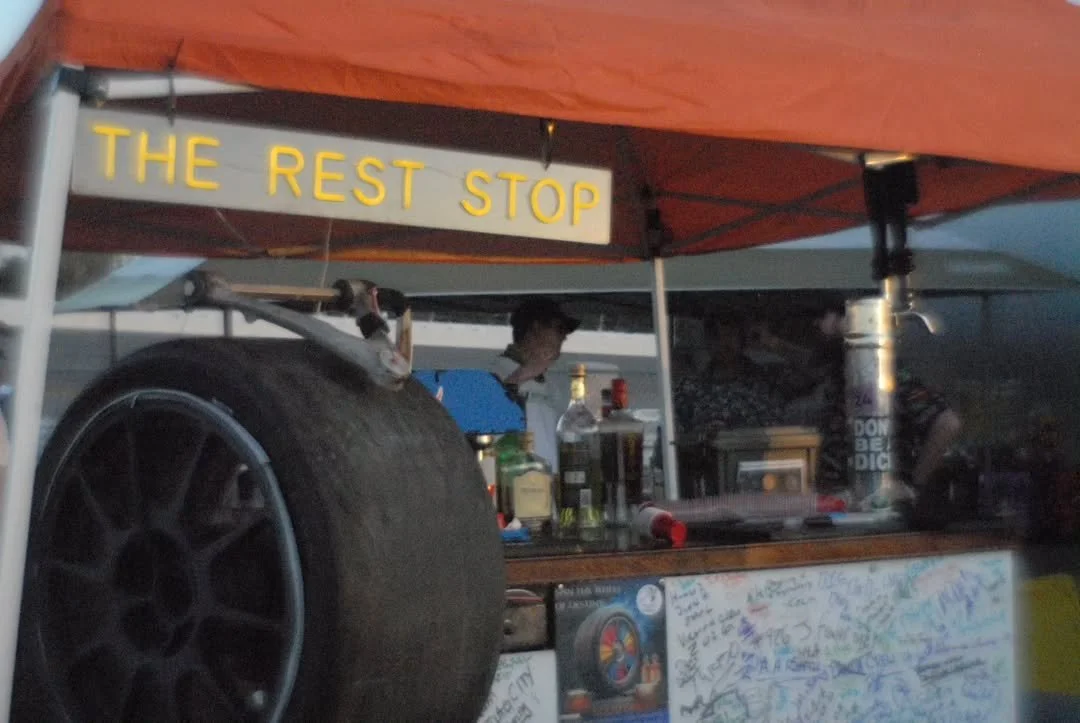 A food or drink stand called 'The Rest Stop' with bottles, a large tire, and a person in the background under an orange canopy.