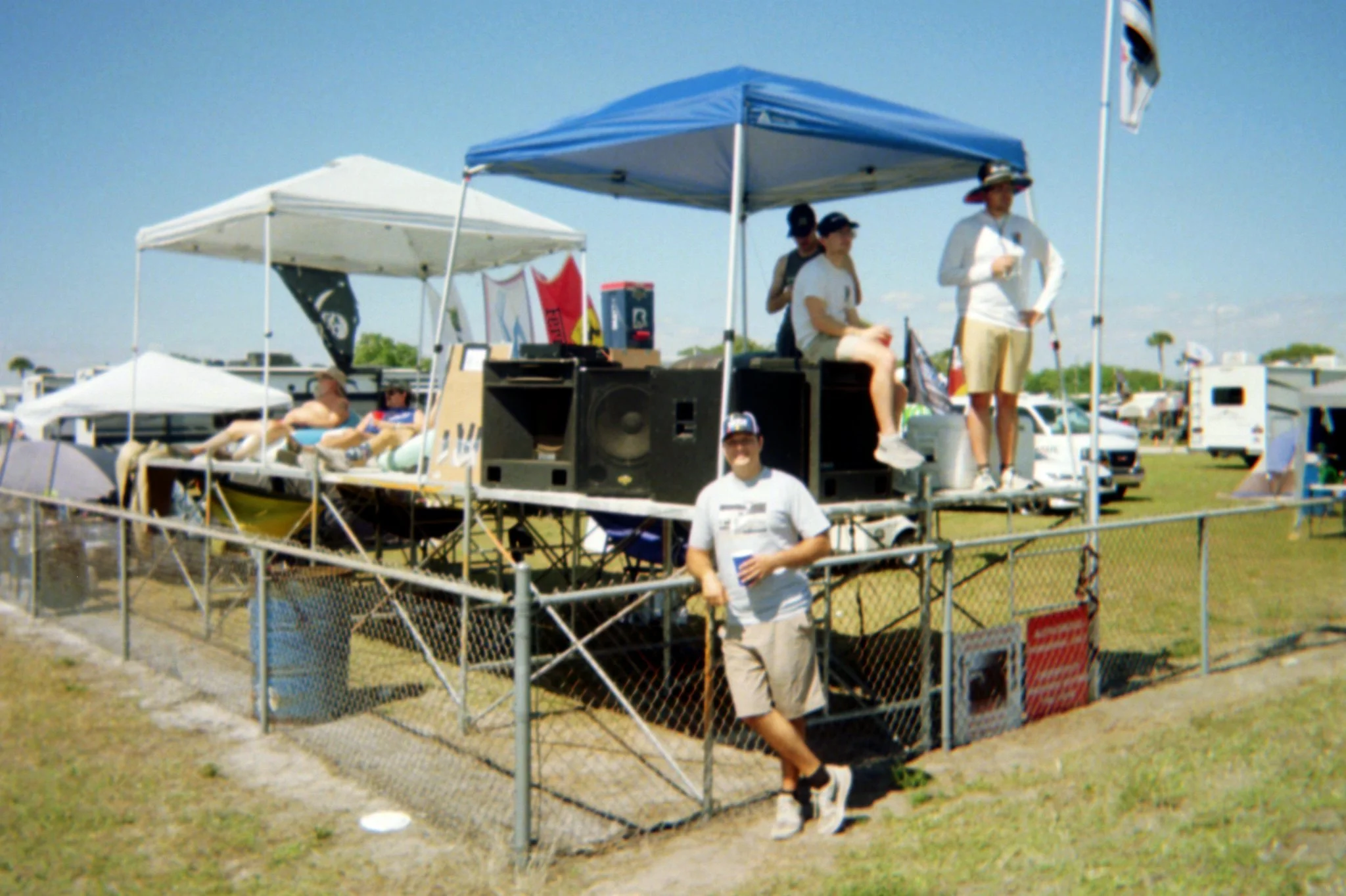 People relaxing on a social gathering platform with tents, speakers, and camping trailers in the background on a sunny day.