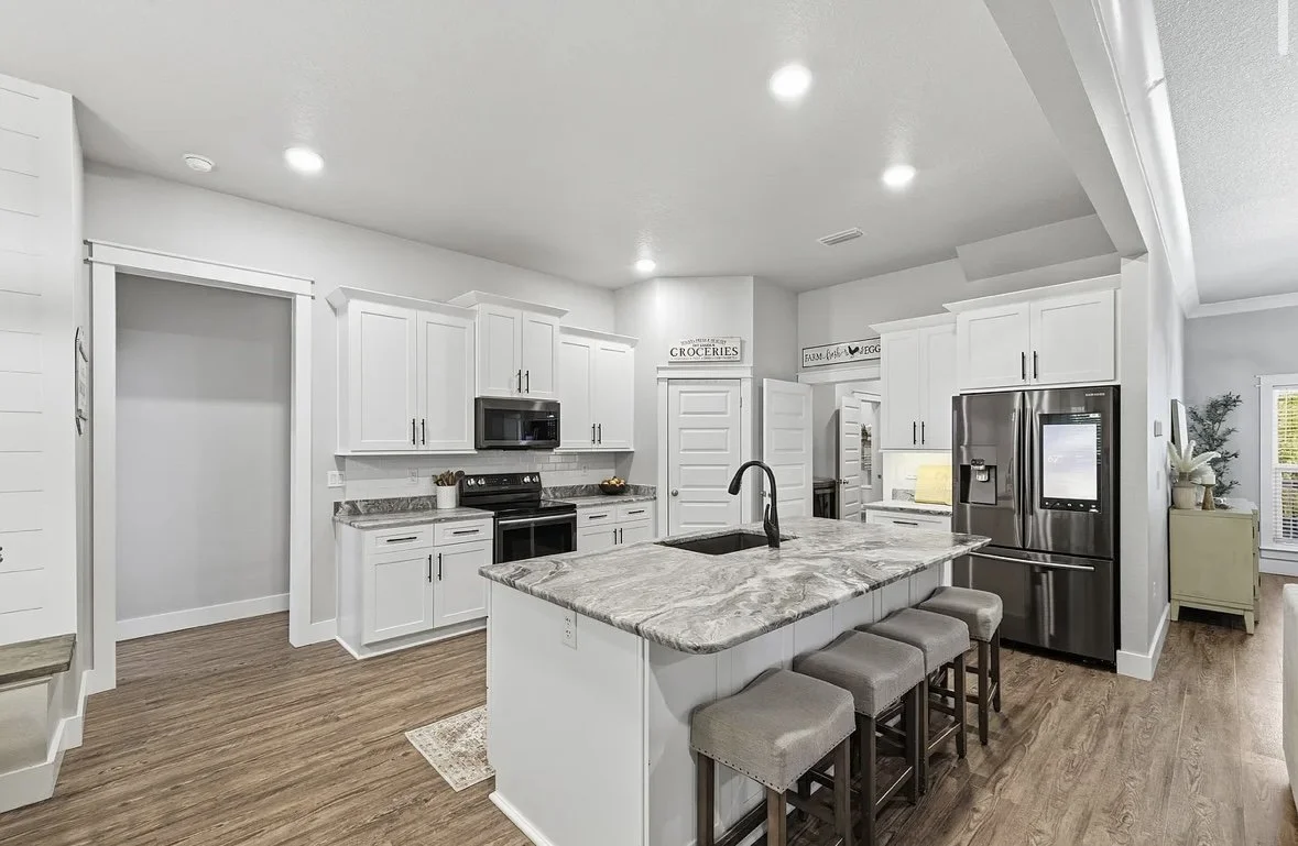 Modern kitchen with white cabinets, granite countertops, stainless steel refrigerator, black stove, microwave, island with sink, and four bar stools. Wooden floors and recessed lighting.