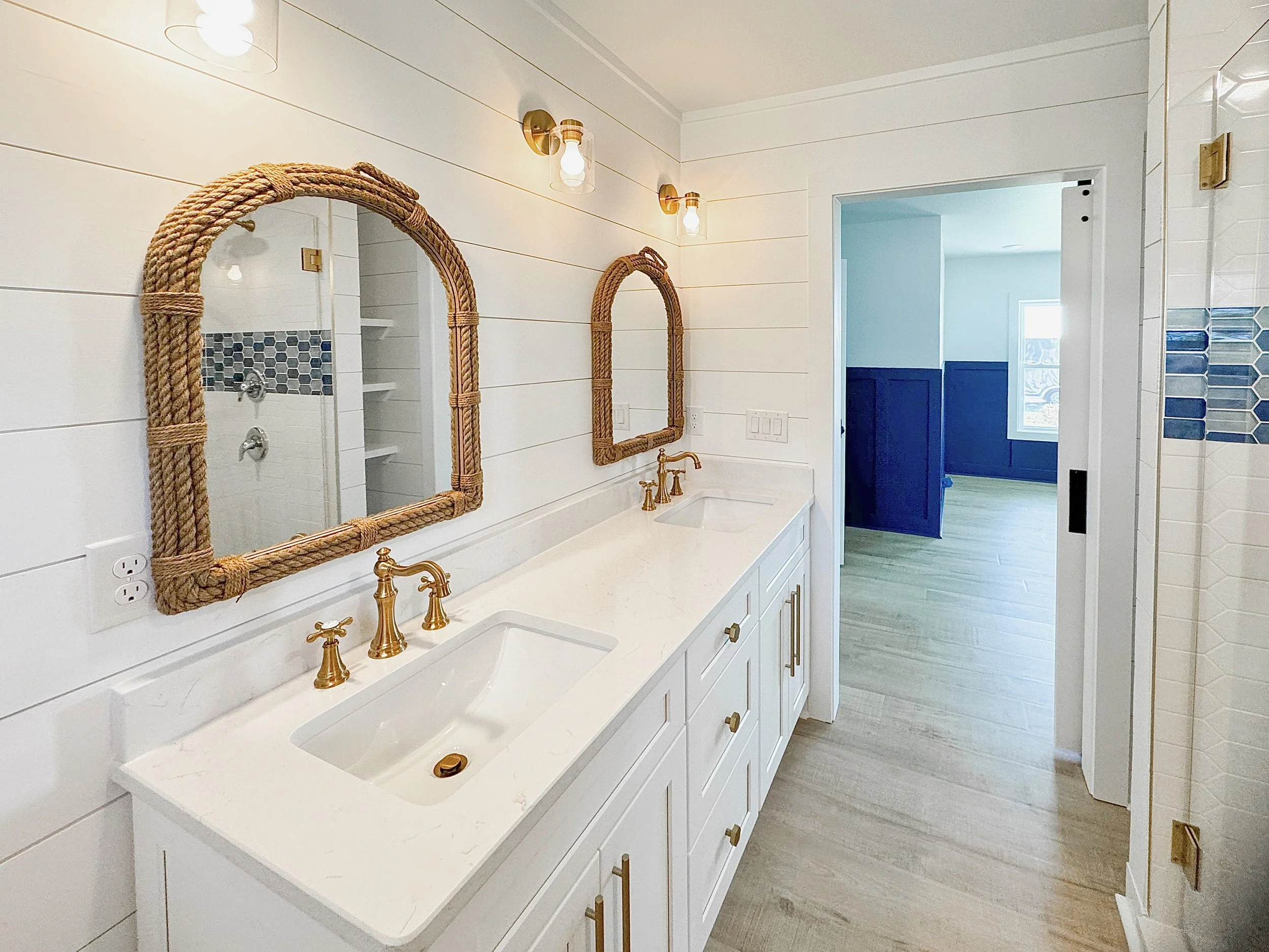 Bathroom with white double vanity, gold fixtures, wicker-framed mirrors, and shiplap walls, leading to a room with blue wainscoting.