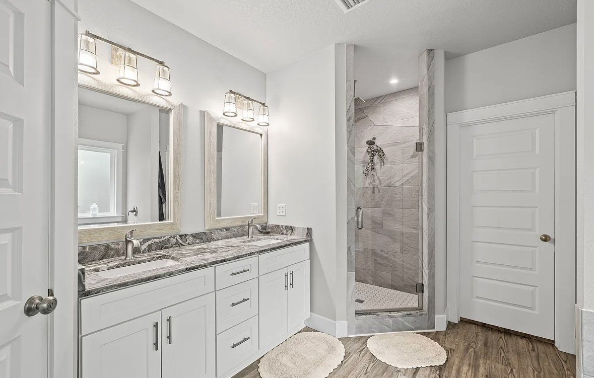 Bathroom with double vanity, marble countertop, two mirrors, modern lighting, walk-in shower with glass door, tiled walls, and wooden flooring.