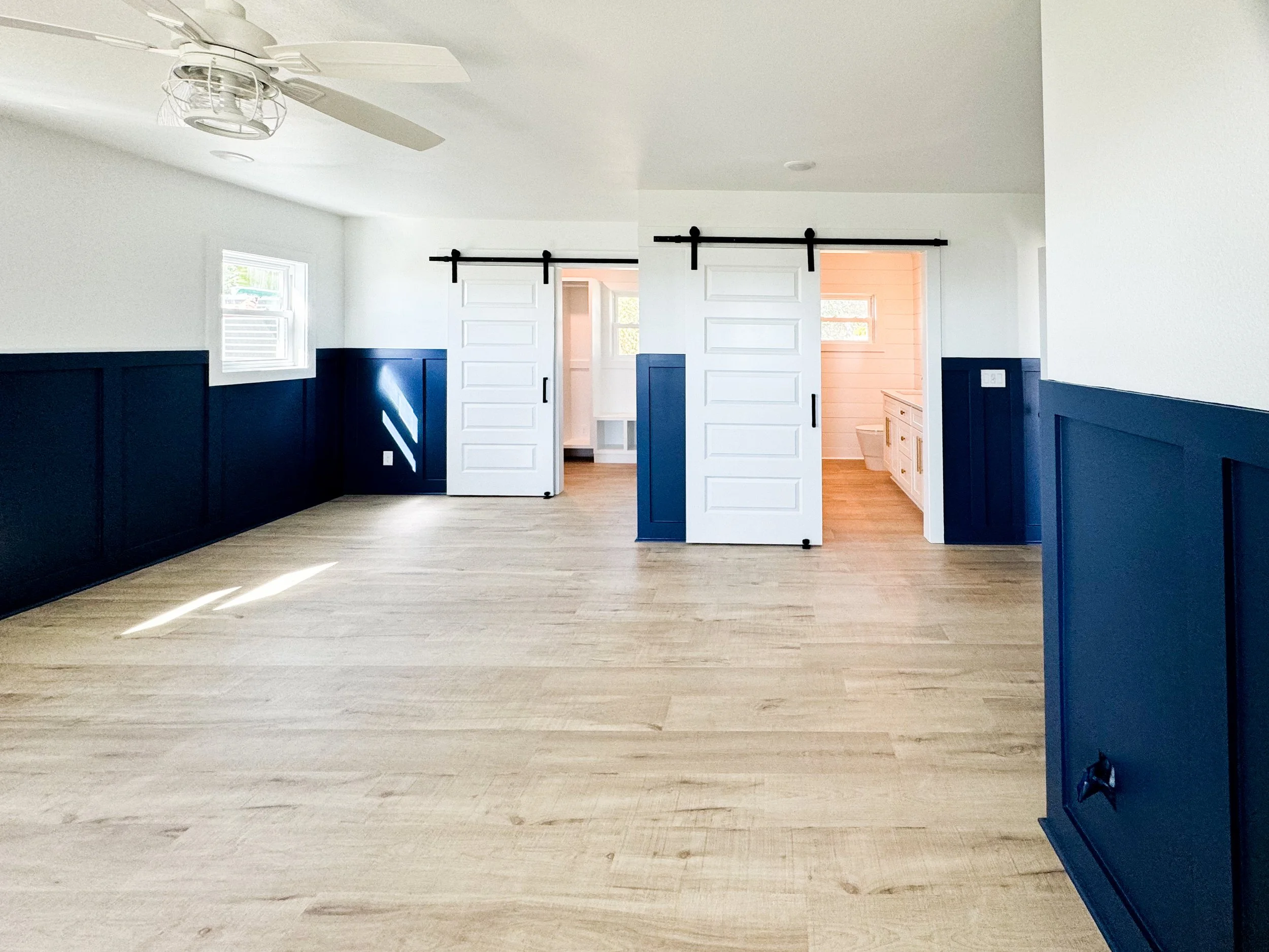 Empty living space with light wood floors, white walls with dark blue wainscoting, a ceiling fan, and two sliding barn doors leading to a bathroom area with a white cabinet and a toilet.