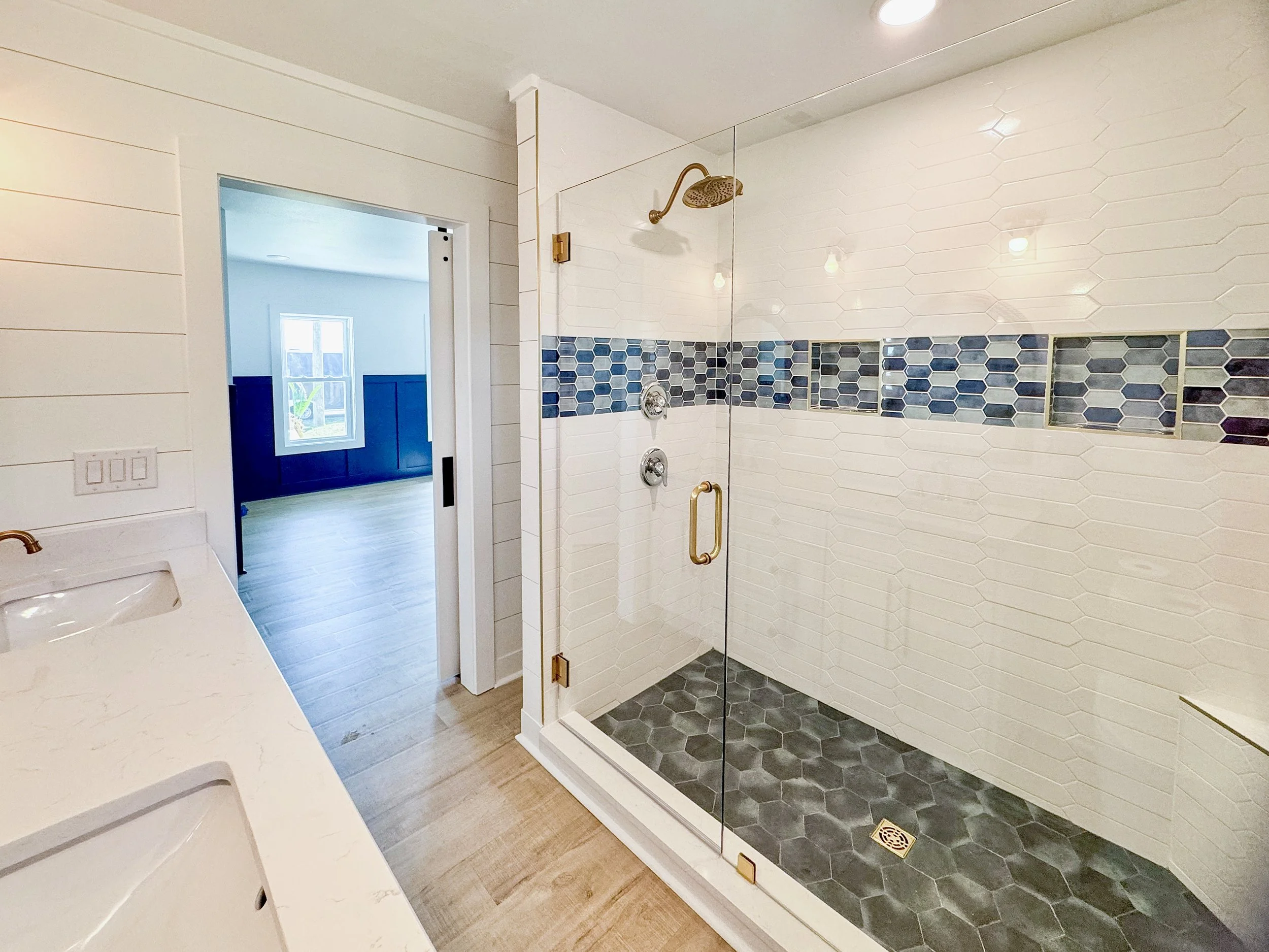 Modern walk-in shower with white hexagon and blue tile accents, glass door, gold fixtures, adjacent double vanity with white countertop, in a bathroom with wood flooring and views into a bedroom with blue wainscoting and a window.