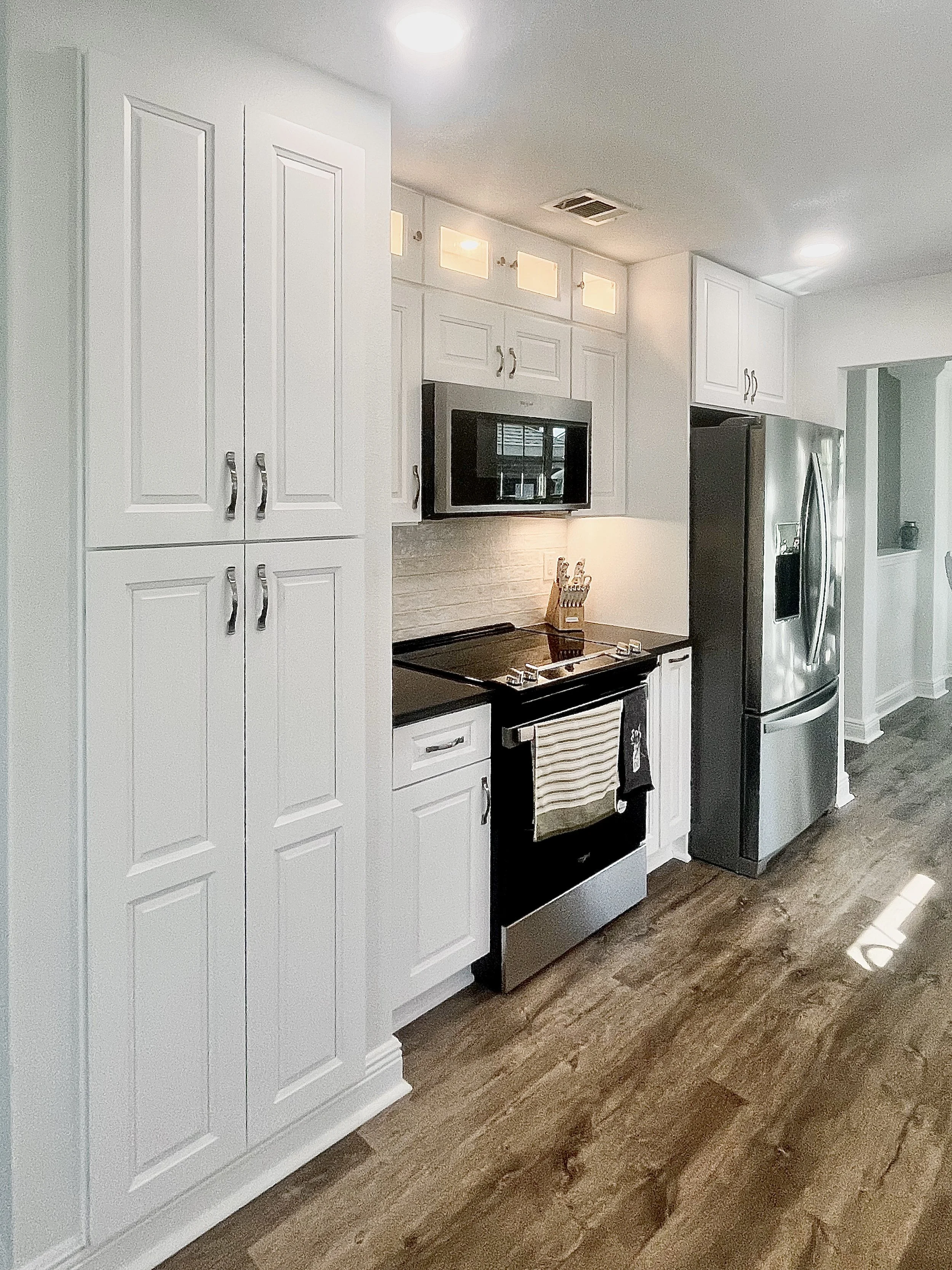 Modern kitchen with white cabinets, stainless steel refrigerator, oven, microwave, and wooden flooring.