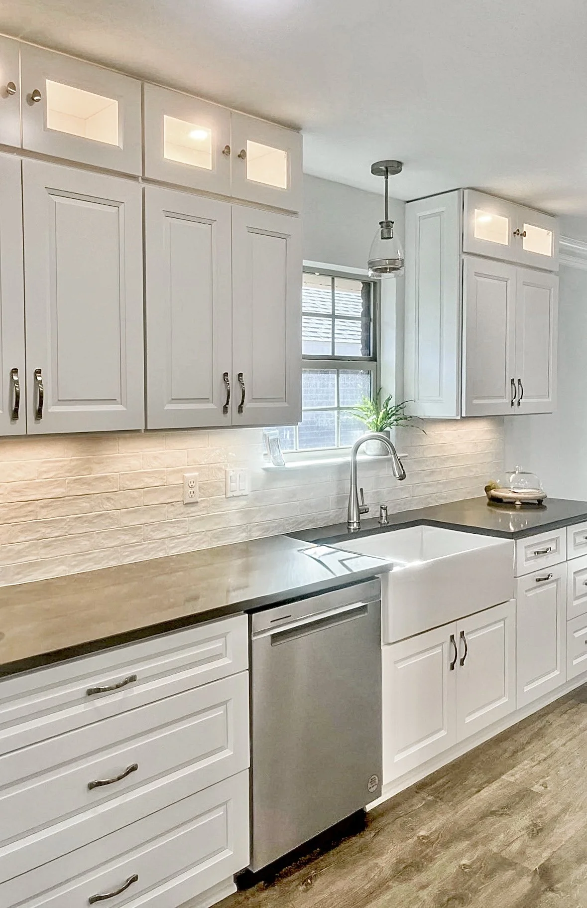 Modern kitchen with white cabinets, a farmhouse sink, a window behind the sink, a potted plant on the window sill, a stainless steel dishwasher, granite countertops, and a rustic wood floor.