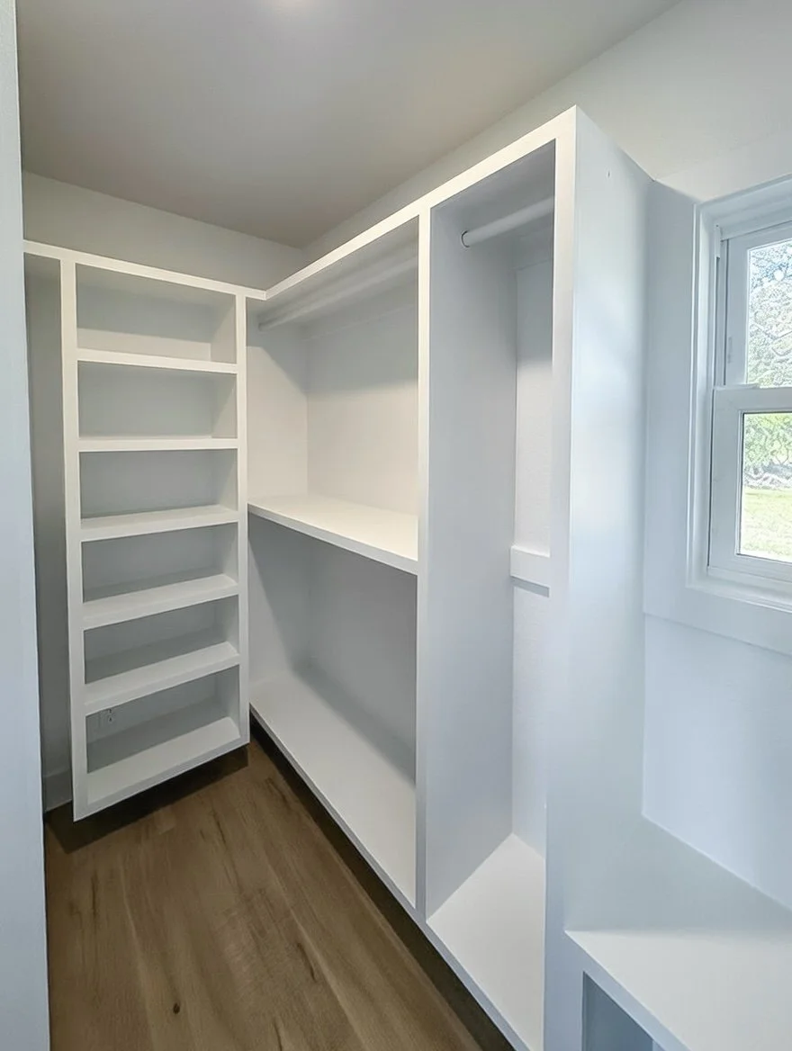 Empty built-in white closet with shelves and hanging rod, located next to a window with a view of trees.