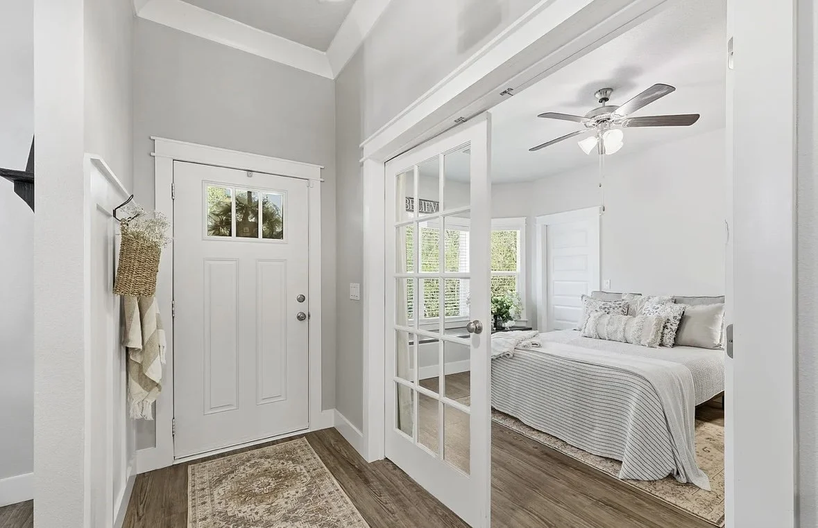 View of a bright, white bedroom seen through a glass-paneled door from an entryway with a front door and wall hanging towels.