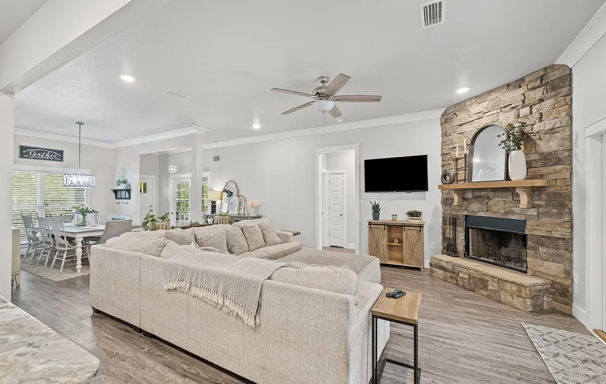 Living room with a beige sectional sofa, wood fireplace with stone surround, wall-mounted TV, and a ceiling fan, adjacent to a dining area with a white table and chairs.