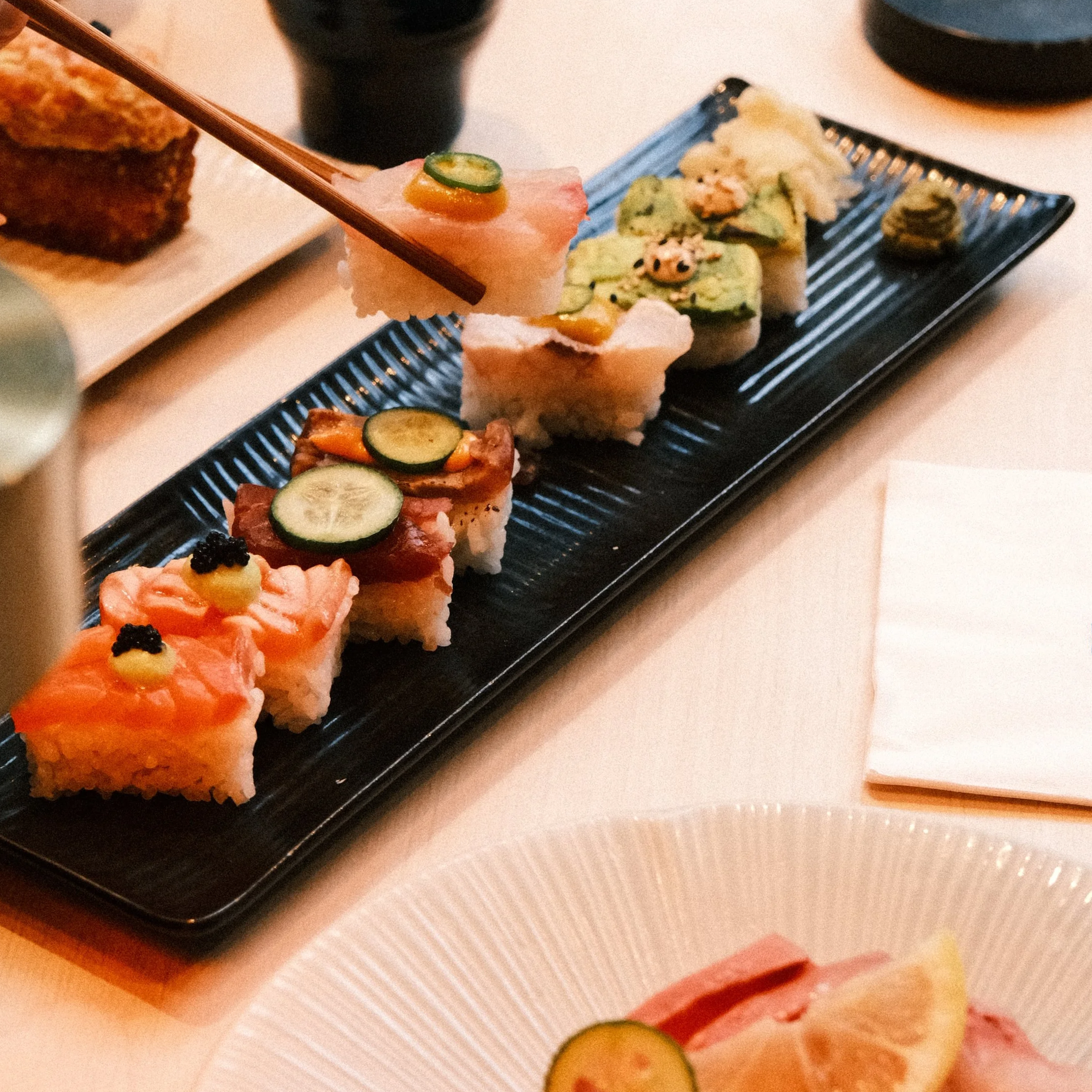 A black rectangular plate with assorted sushi rolls topped with cucumber and fish roe, set on a white table with a white napkin and a plate of sliced lemon and cold cuts in the foreground.