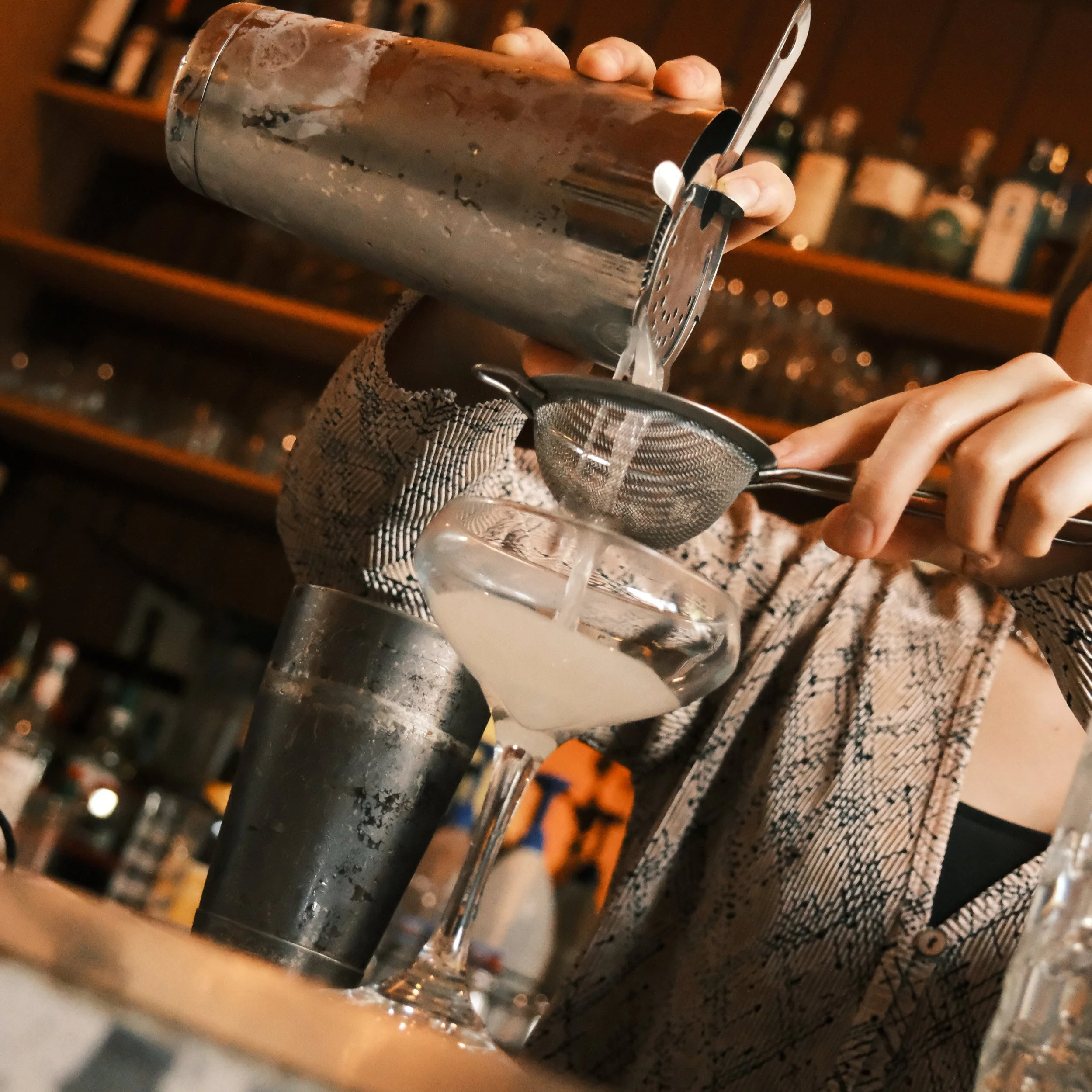A bartender skillfully pouring a cocktail through a strainer into a coupe glass, with blurred bottles in the background, creating a cozy ambiance.