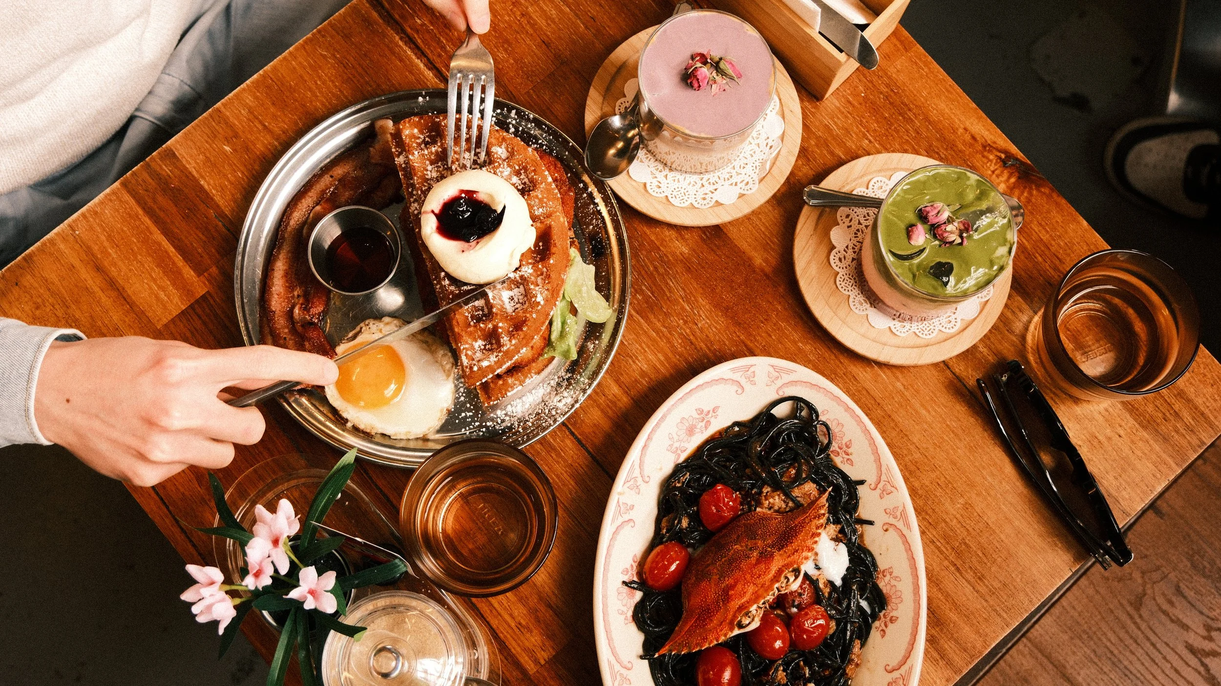 A meal on a wooden table featuring waffles with a fried egg, whipped cream, and berry sauce, alongside a plate of squid ink pasta with cherry tomatoes, two colorful drinks garnished with rose petals, glasses of water, and a small vase with pink flowers.