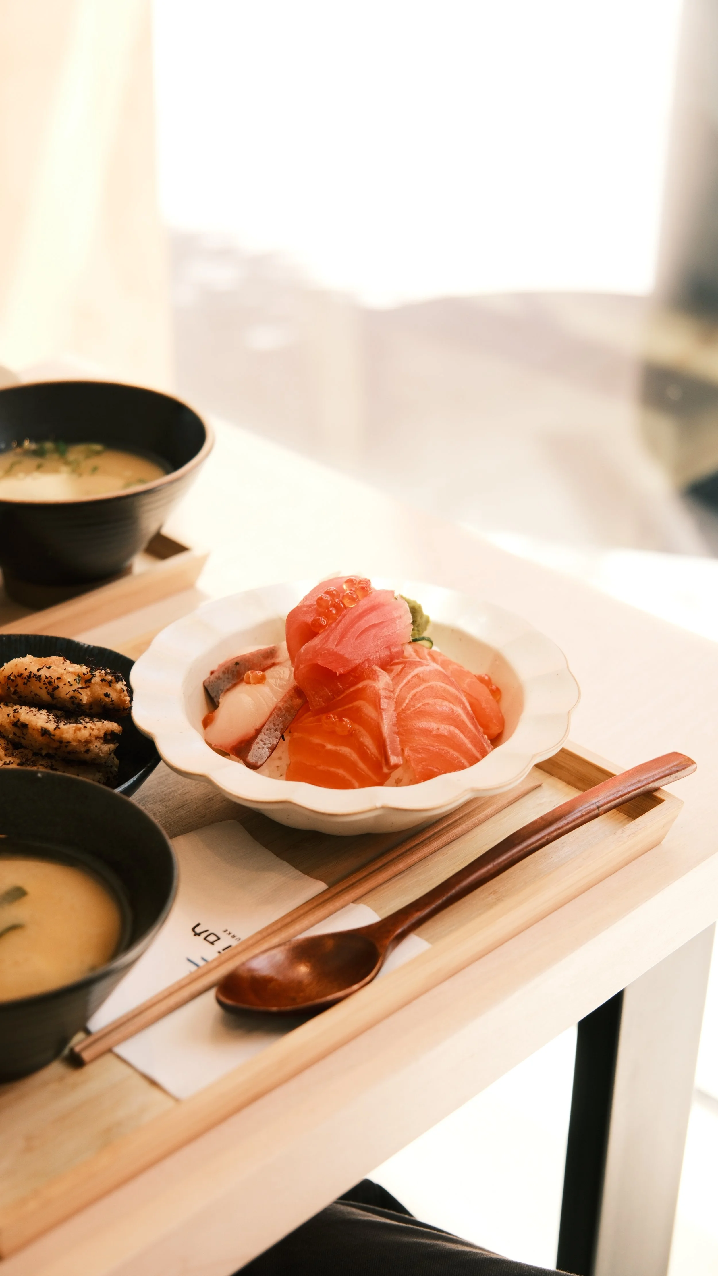 A table setting with a white bowl of assorted sashimi, featuring pink and orange fish slices, next to miso soup and tempura, on a wooden tray.