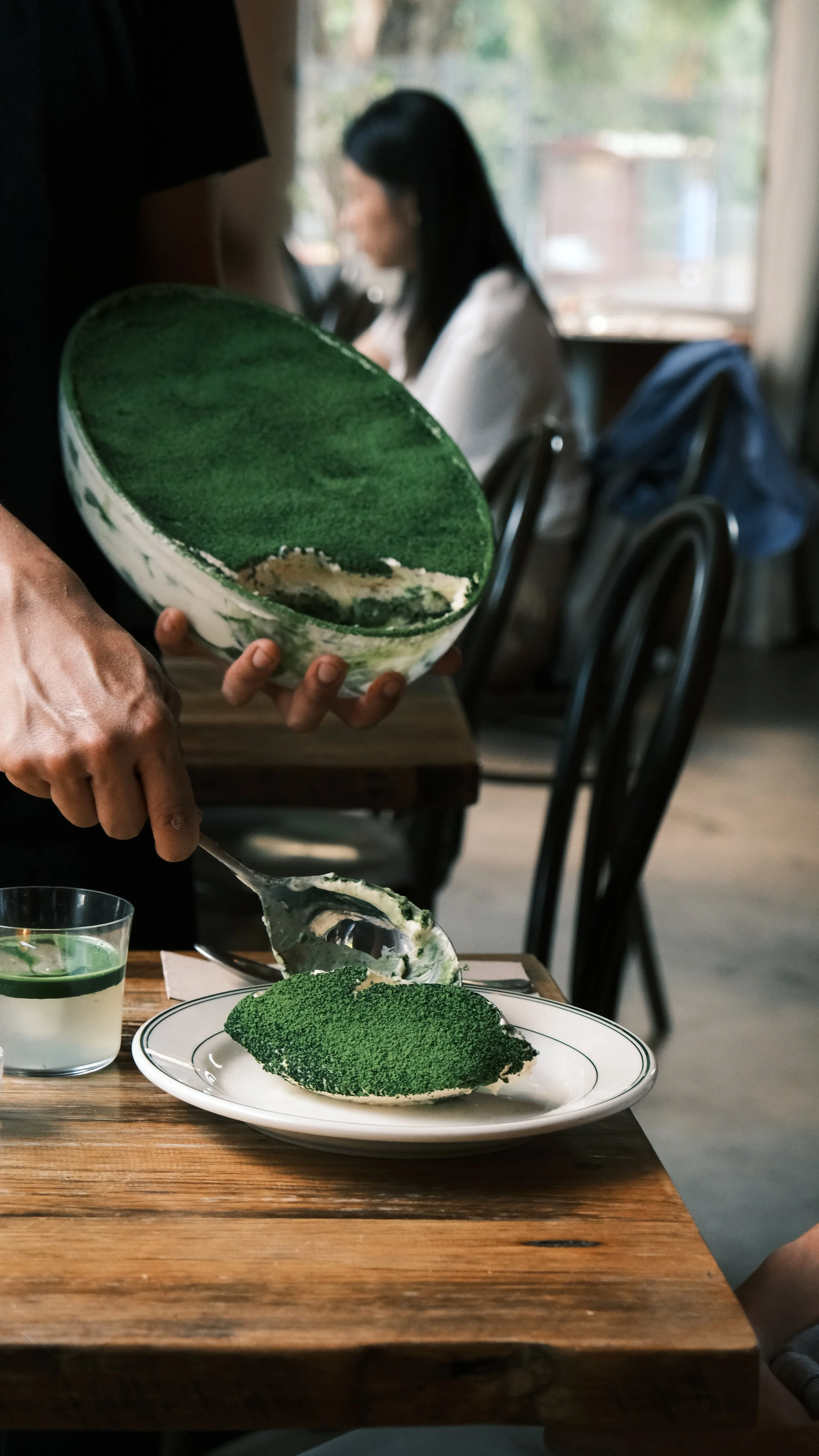Person serving a slice of matcha cake at a dining table, with a woman sitting in the background near a window.