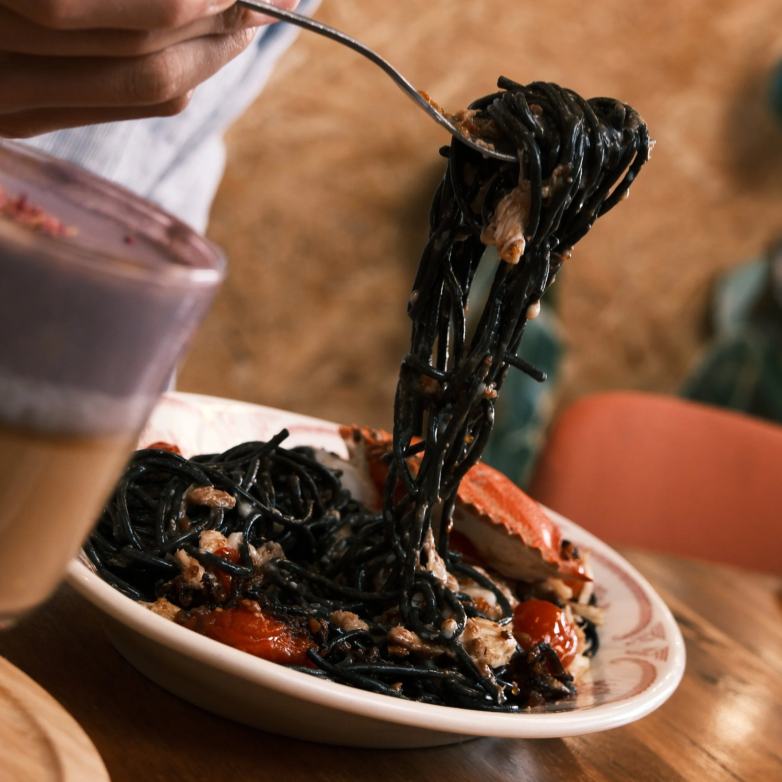 Close-up of a person lifting a forkful of black squid ink noodles with seafood and cherry tomatoes on a plate, with a drink in the foreground.