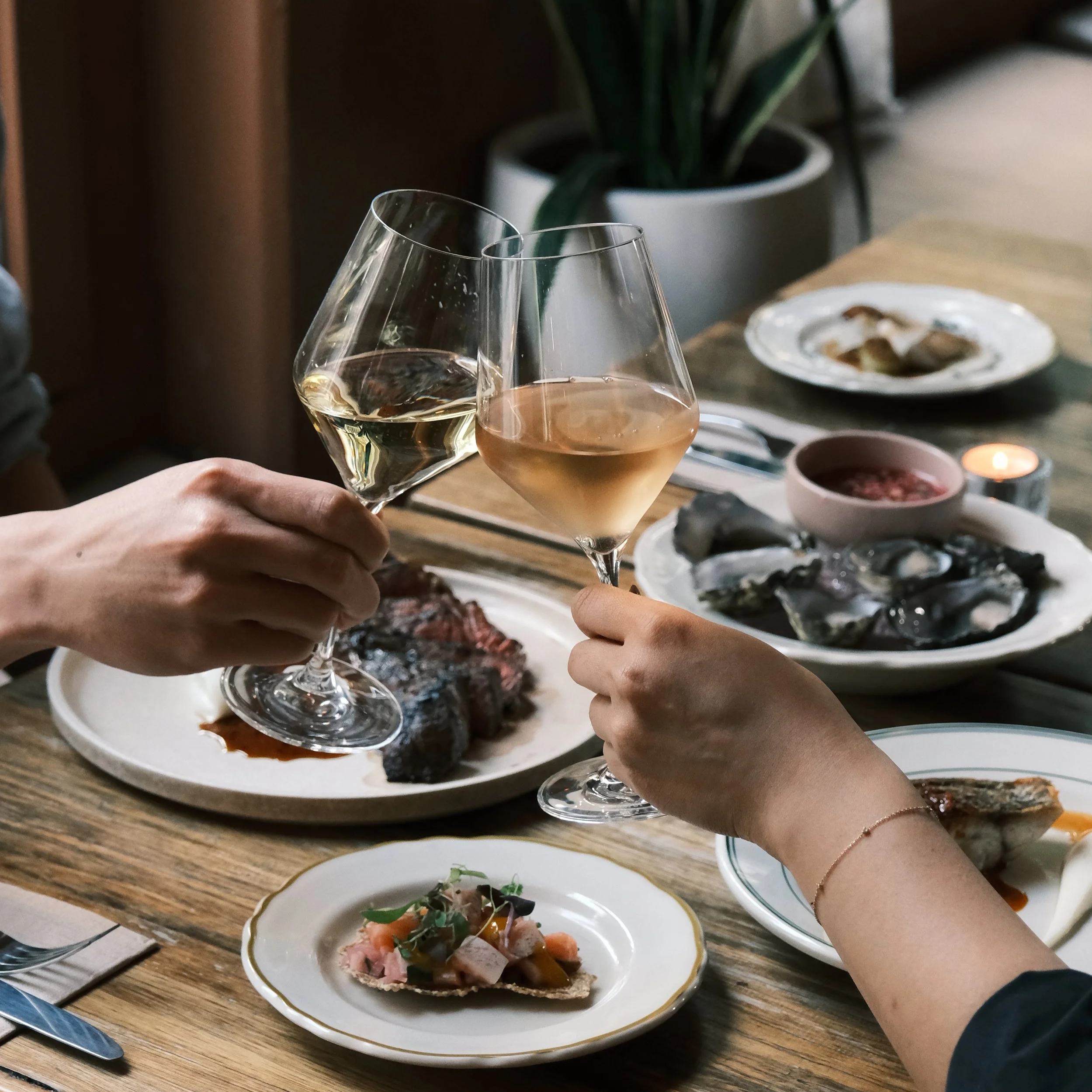 Two people clinking glasses of white wine at a dinner table with various dishes, including oysters, sliced meat, and appetizers.