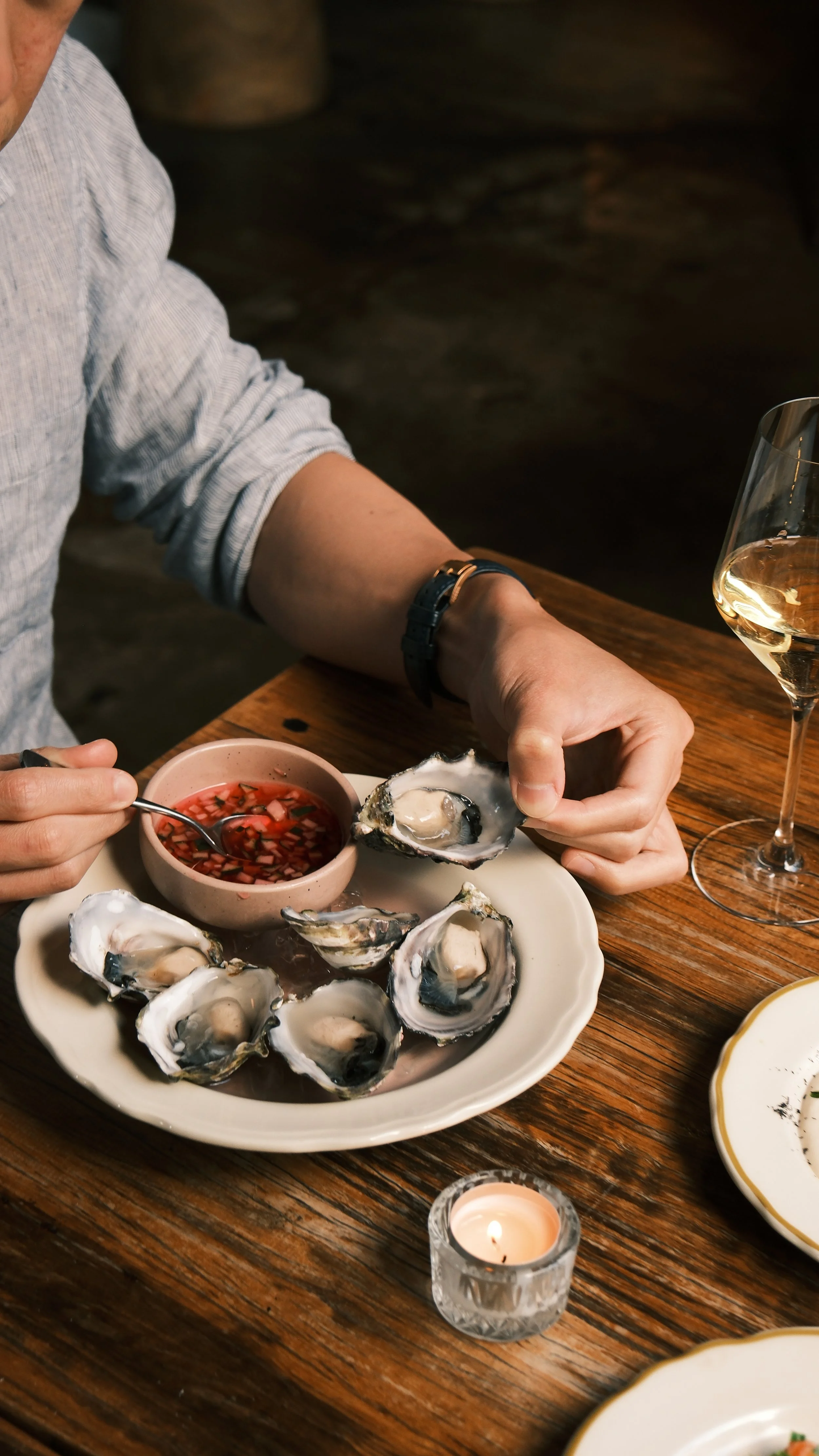 Person holding a freshly shucked oyster, with a small bowl of cocktail sauce and a glass of white wine on a wooden table.