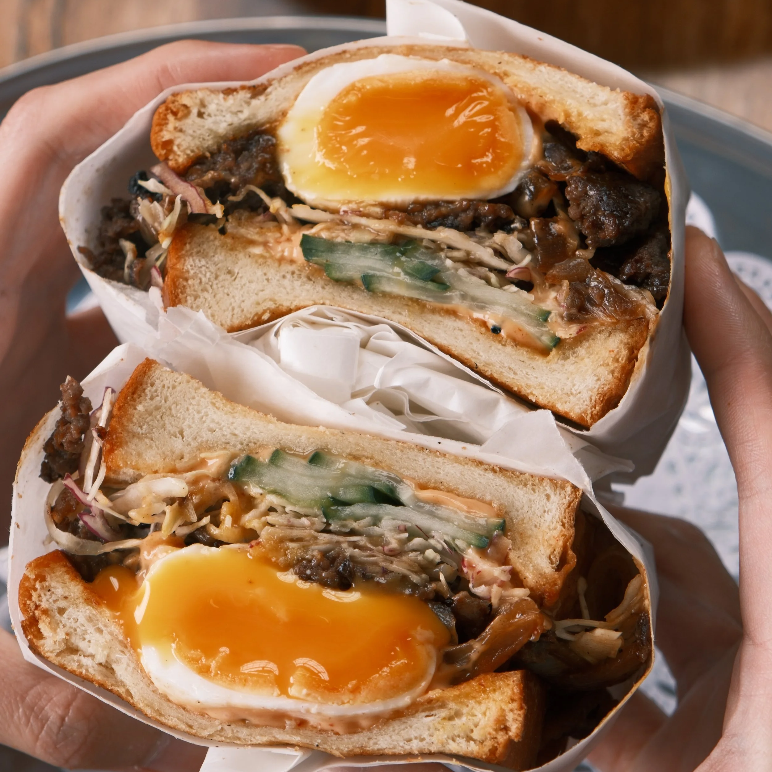 A close-up view of a sandwich with soft white bread, filled with an egg cooked soft with a runny yolk, shredded meat, vegetables, and sauce.