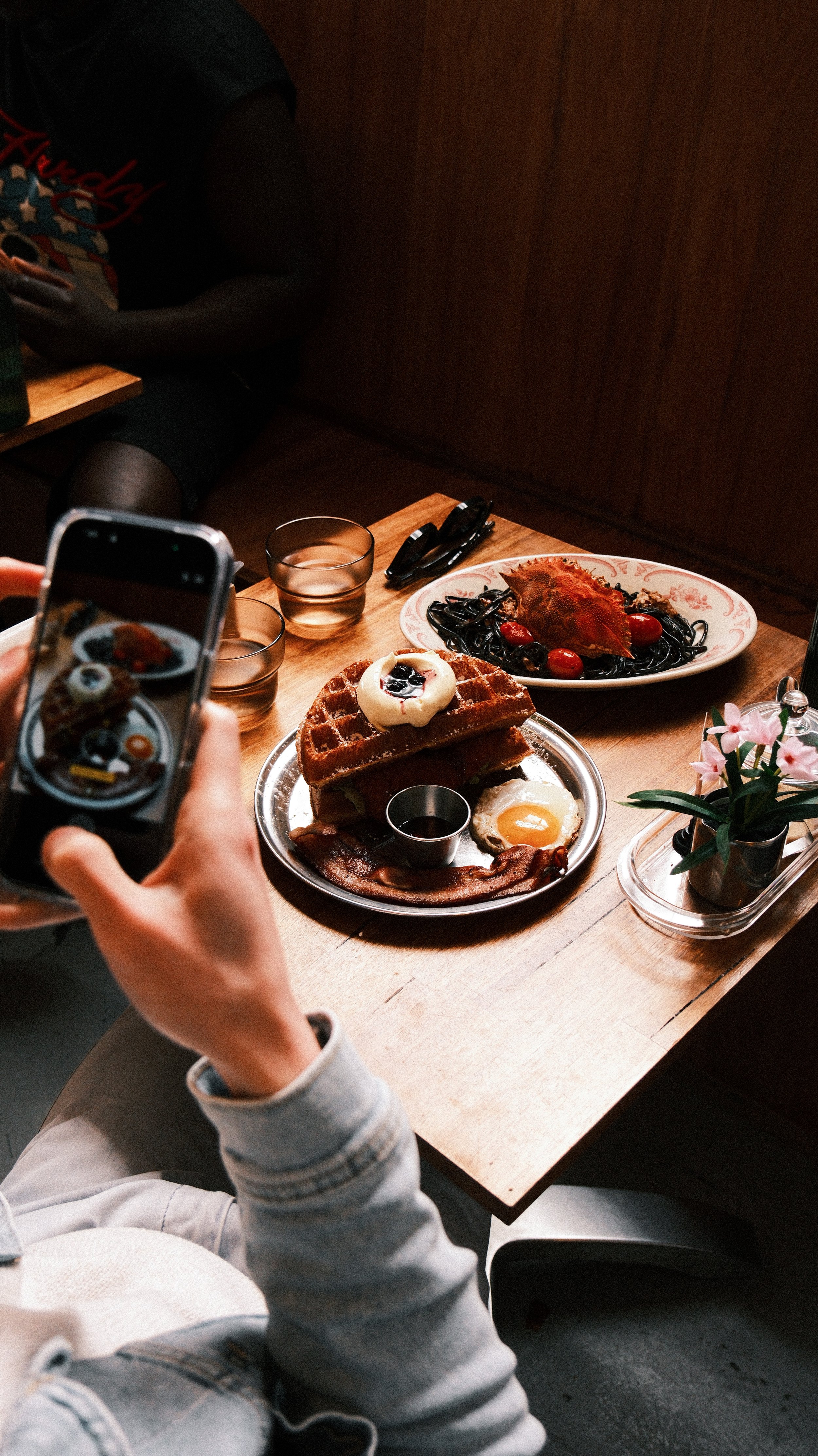 A cozy cafe scene with waffles topped with cream and berries, a fried egg, and sausages. A person photographs the meal, conveying a warm, inviting atmosphere.