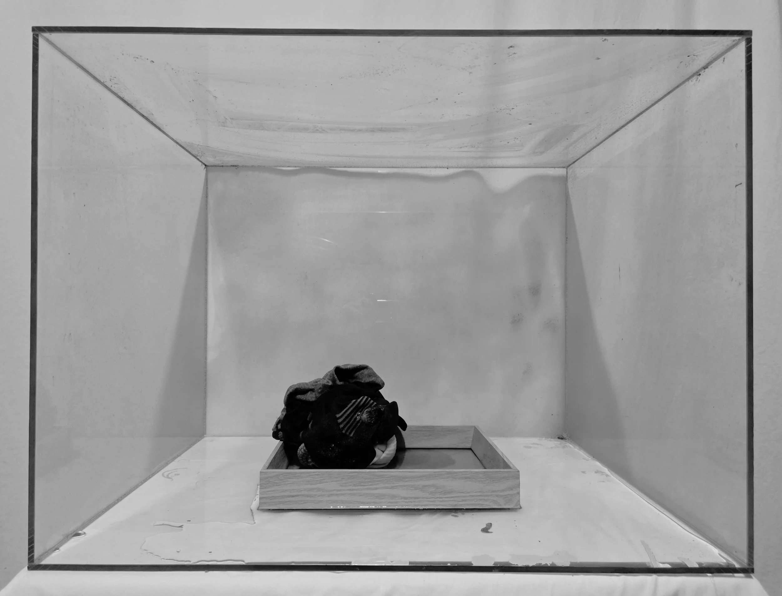 Small pile of clothes and a bag inside a wooden tray, placed inside a box with plain walls.