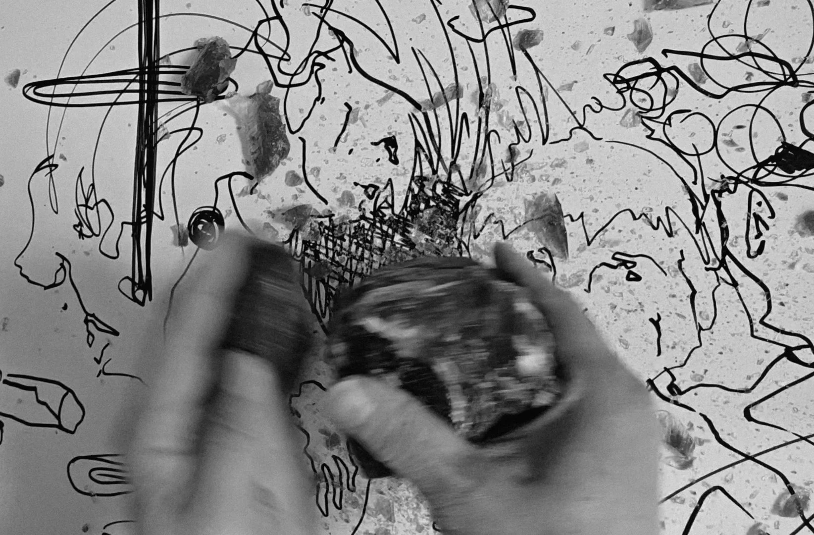 A person with gloved hands drawing a sketch of a horse on a tabletop with a black marker. The background has splattered paint and scattered paper pieces.