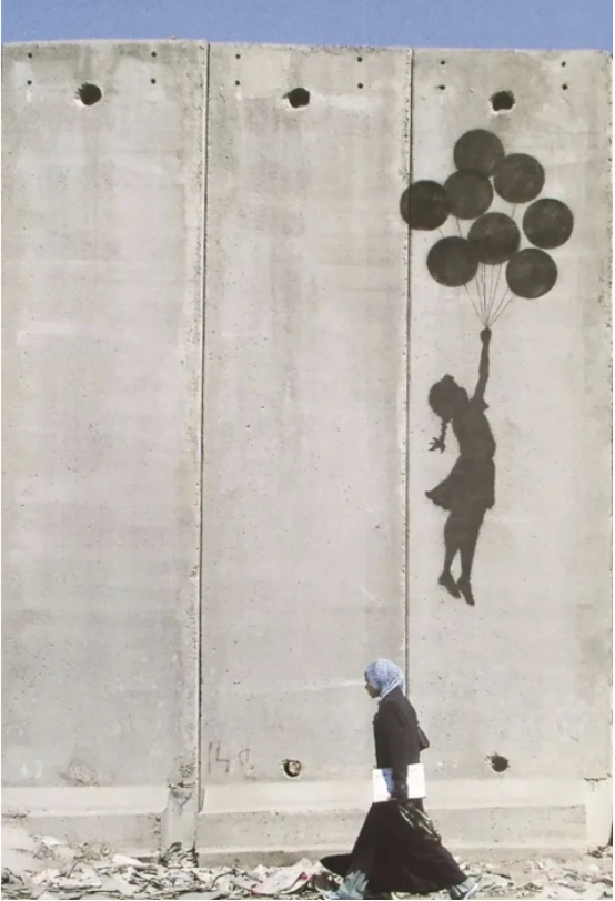 Street art of a girl holding black balloons painted on a concrete wall, with a person walking in front of it.