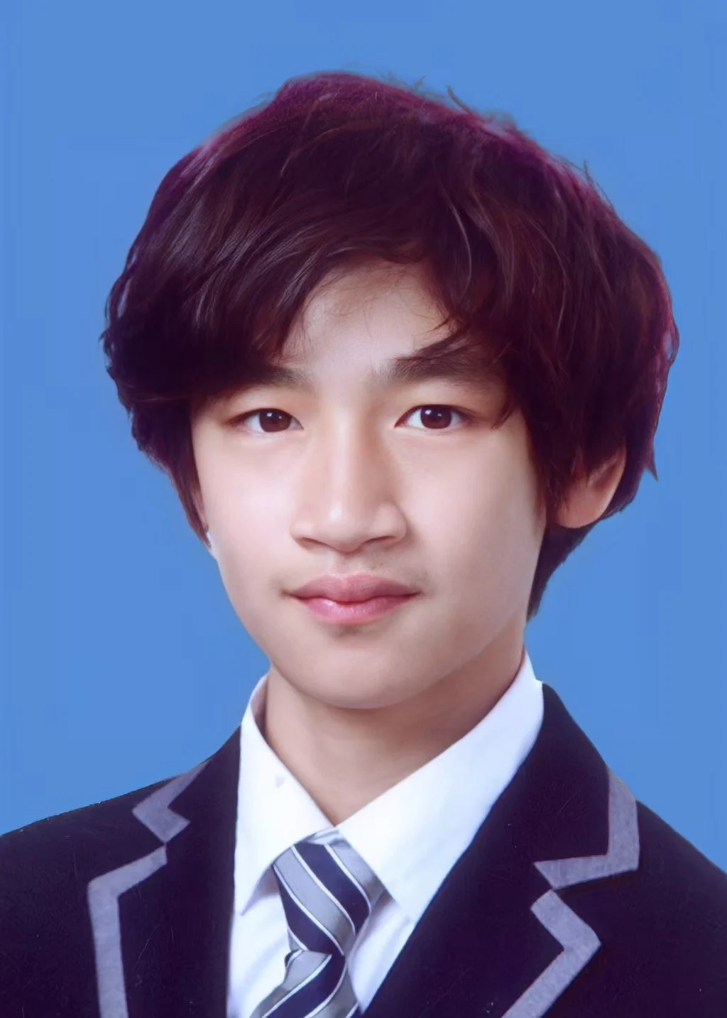 A young man with wavy brown hair, wearing a black blazer with gray trim, a white shirt, and a striped tie, against a blue background.