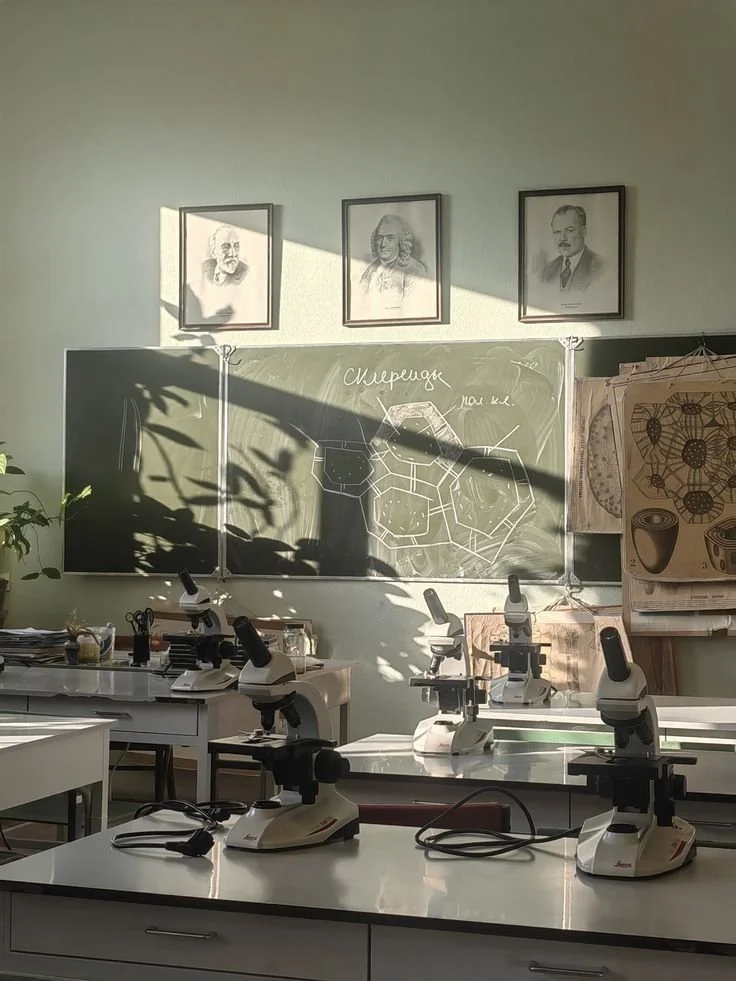 Science classroom with microscopes on tables, framed portraits above blackboard, botanical illustrations on the wall, and sunlight casting shadows.