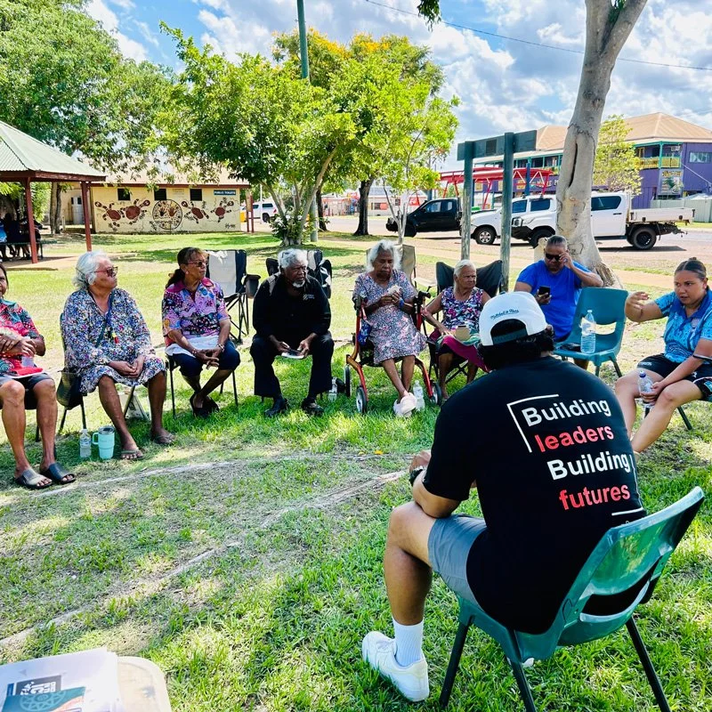 A group of elderly women and a few younger people sitting outdoors in a semi-circle on green grass, participating in a community gathering under trees with a building and parked cars in the background. One person in the foreground wears a black t-shirt with the words 'Building leaders Building futures' on the back.