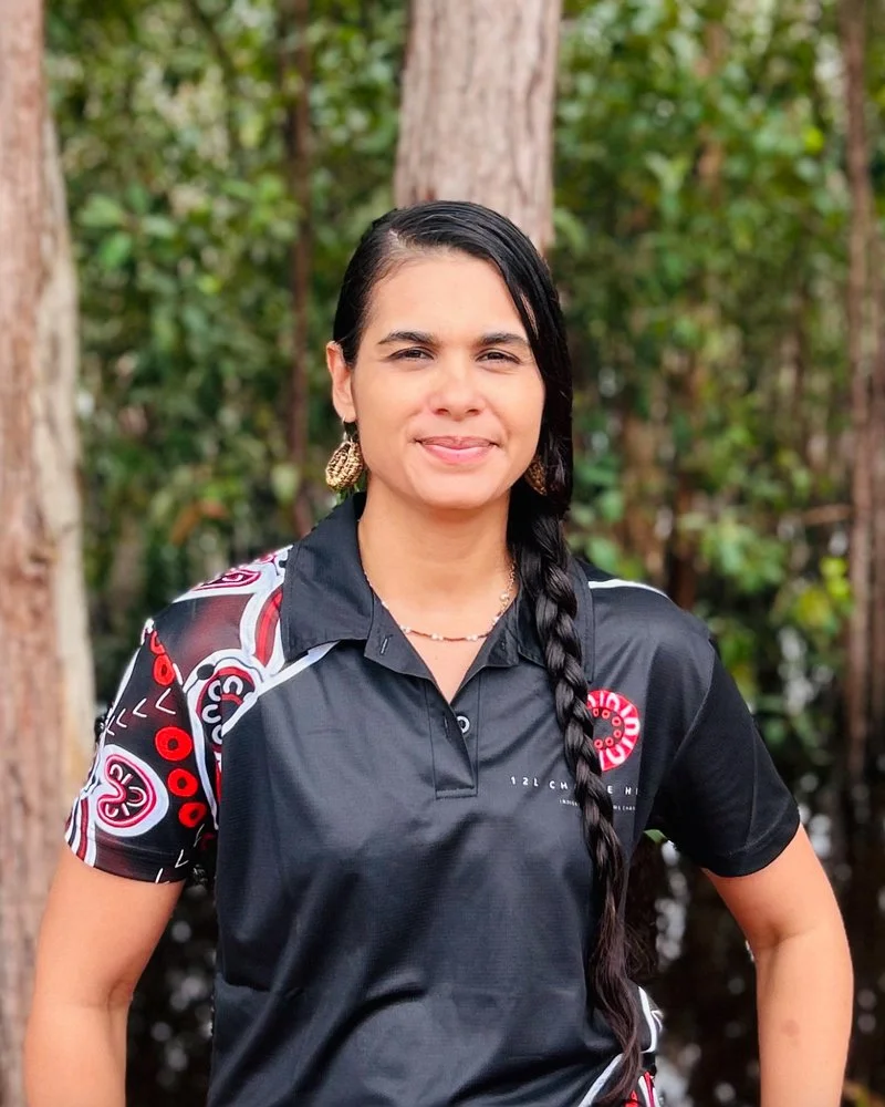 A woman with long dark hair in a braid standing outdoors with trees in the background. She is wearing a black sports shirt with colorful patterns and logo, earrings, and a necklace, smiling slightly.