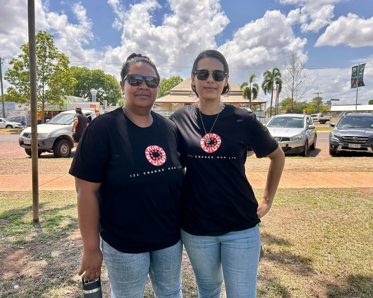 Two women standing outdoors wearing matching black T-shirts with the I2L Change Hub logo, both wearing sunglasses, with cars and trees in the background under a partly cloudy sky.