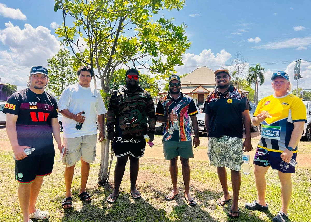 Six men standing outdoors under a tree with green leaves. They are dressed in casual and sportswear, some holding water bottles. The background shows a house, palm trees, parked cars, and a partly cloudy sky.