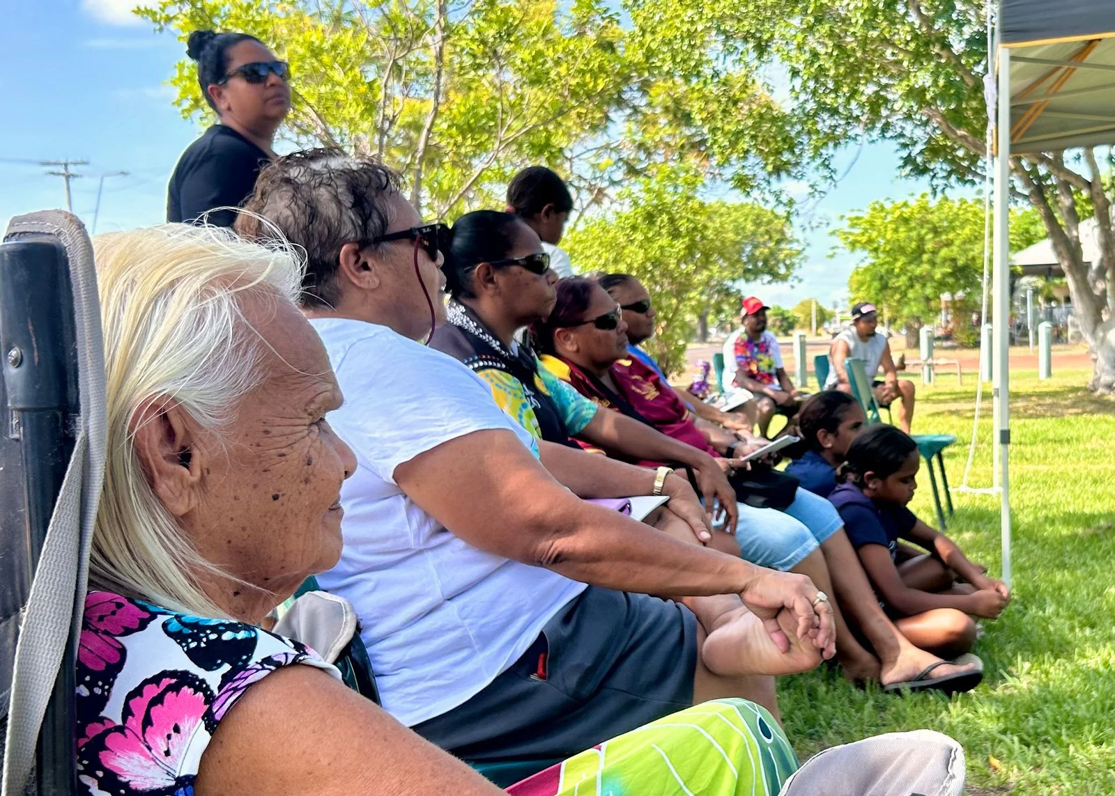 A group of diverse people sitting outdoors on chairs and the ground during daytime, under the shade of trees, attending an event or gathering.