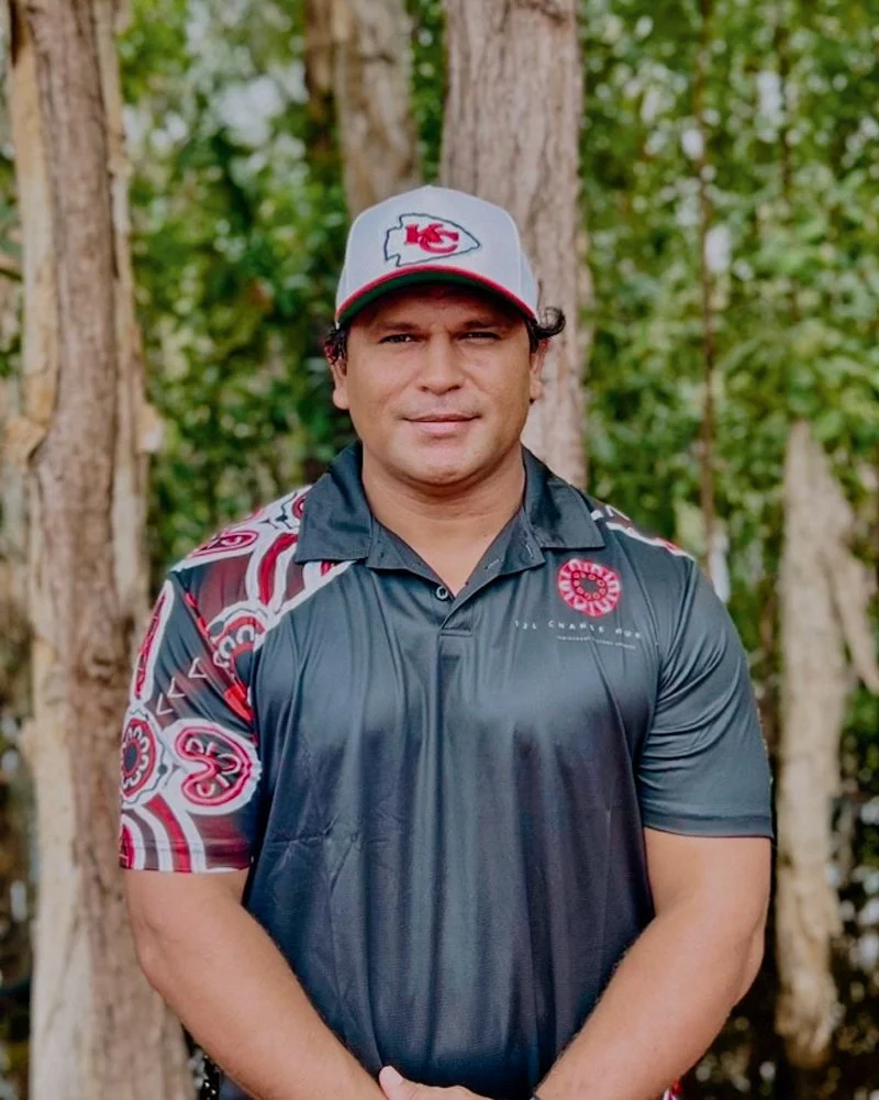 A man wearing a gray cap, a black and red sports polo shirt, standing outdoors in front of trees.