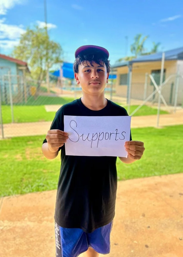 A boy is standing outdoors on a sunny day, holding a paper that says 'Supports,' wearing a black shirt, blue shorts, and a purple cap. The background includes a grassy yard, a house, and a blue sky.