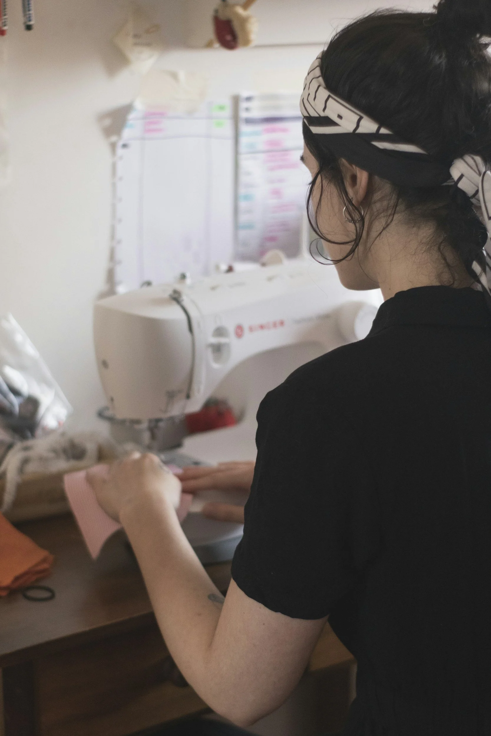 A person wearing a black shirt and a black-and-white headscarf working on a fabric project at a wooden table, with a sewing machine and colorful paper pieces nearby.