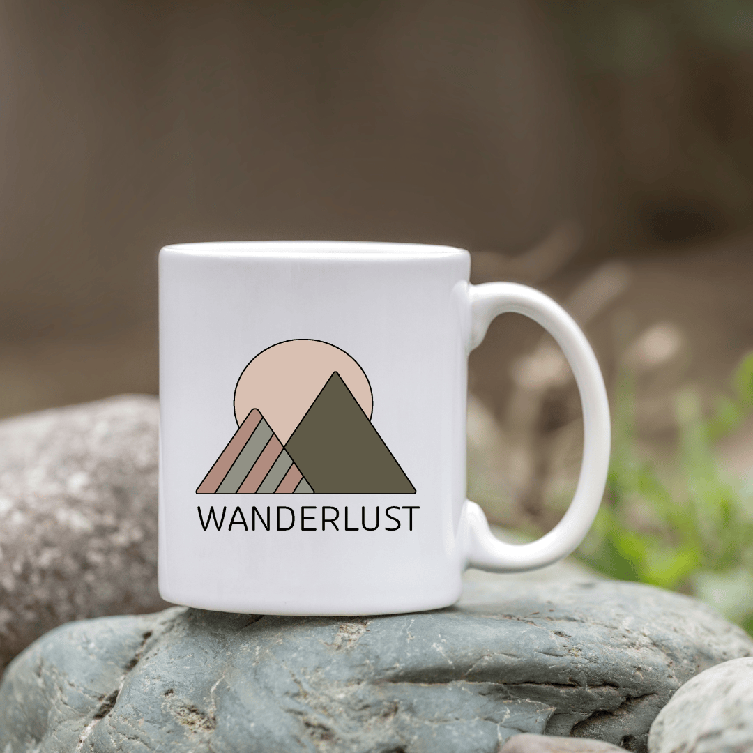 White ceramic mug with a minimalist mountain design and the word ‘WANDERLUST,’ placed on a rock outdoors.