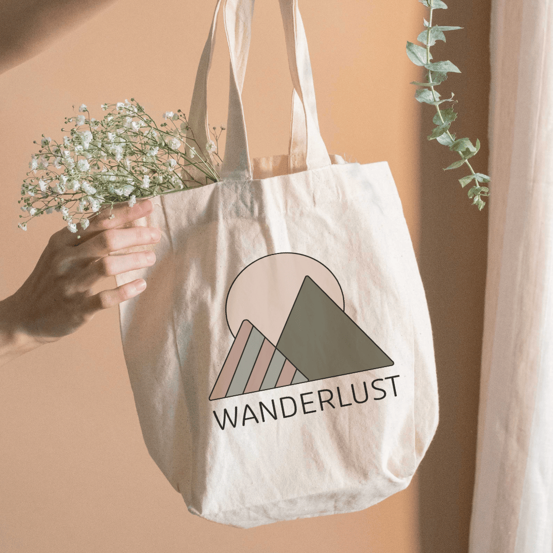 Hand holding a beige tote bag with a minimalist mountain design and the word ‘WANDERLUST,’ filled with small white flowers.