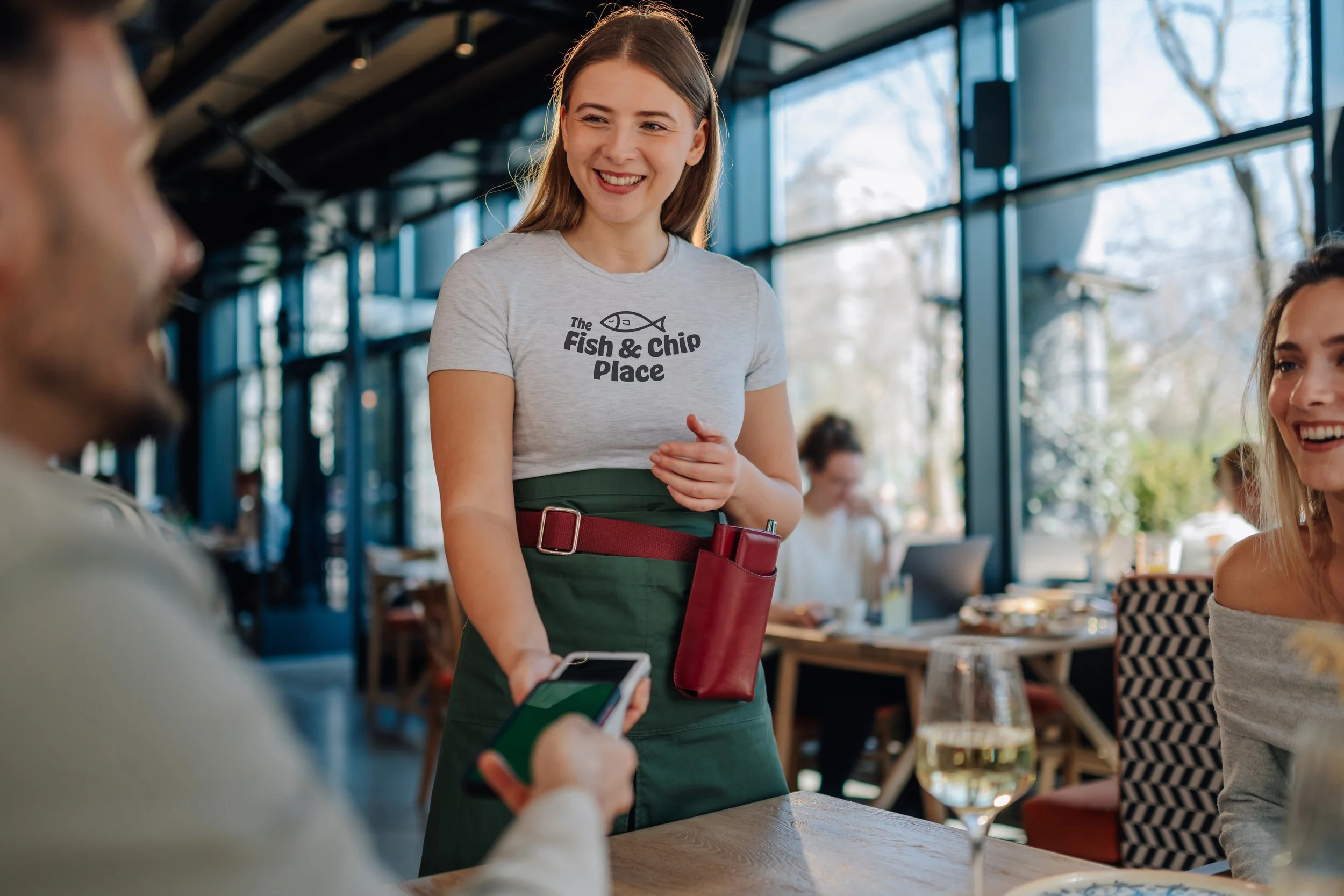Server with restaurant logo on their t-shirt and two people sitting at a table in an indoor restaurant setting.