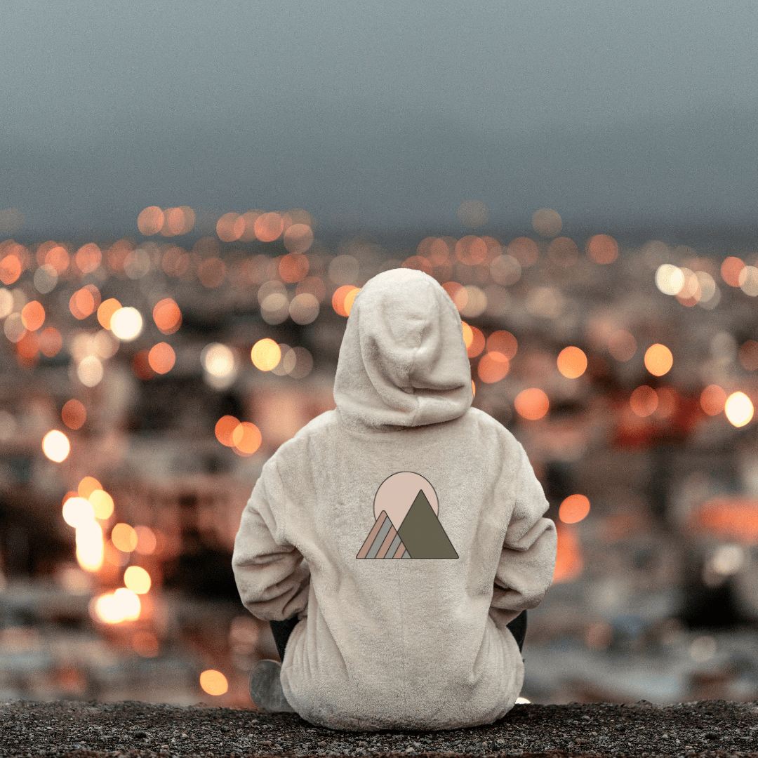Person wearing a hooded sweater, looking out over a blurred cityscape at dusk. The sweater they are wearing has the apparel company's logo that consists of a simple mountain and sun graphic.
