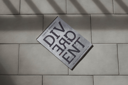 A paperback copy of “Divergent” lies diagonally on a tiled floor, its bold black title split across three stacked lines. Soft shadows from vertical slats fall across the cover and floor, creating a dramatic, slightly confined atmosphere.
