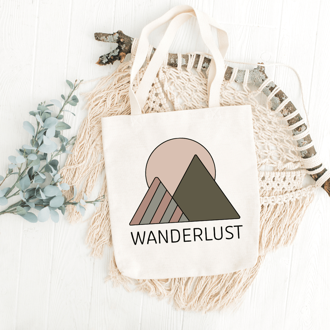 Beige canvas tote bag with a minimalist mountain design and the word ‘WANDERLUST,’ placed on a white wooden surface.