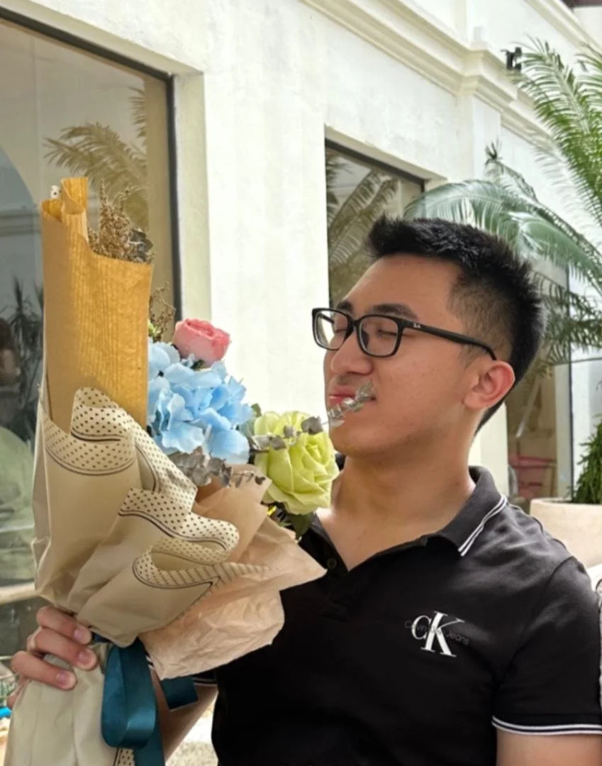 A man with black glasses and a black polo shirt holding a bouquet of colorful flowers, including pink, yellow, and blue, indoors with a white wall, windows, and green plants in the background.
