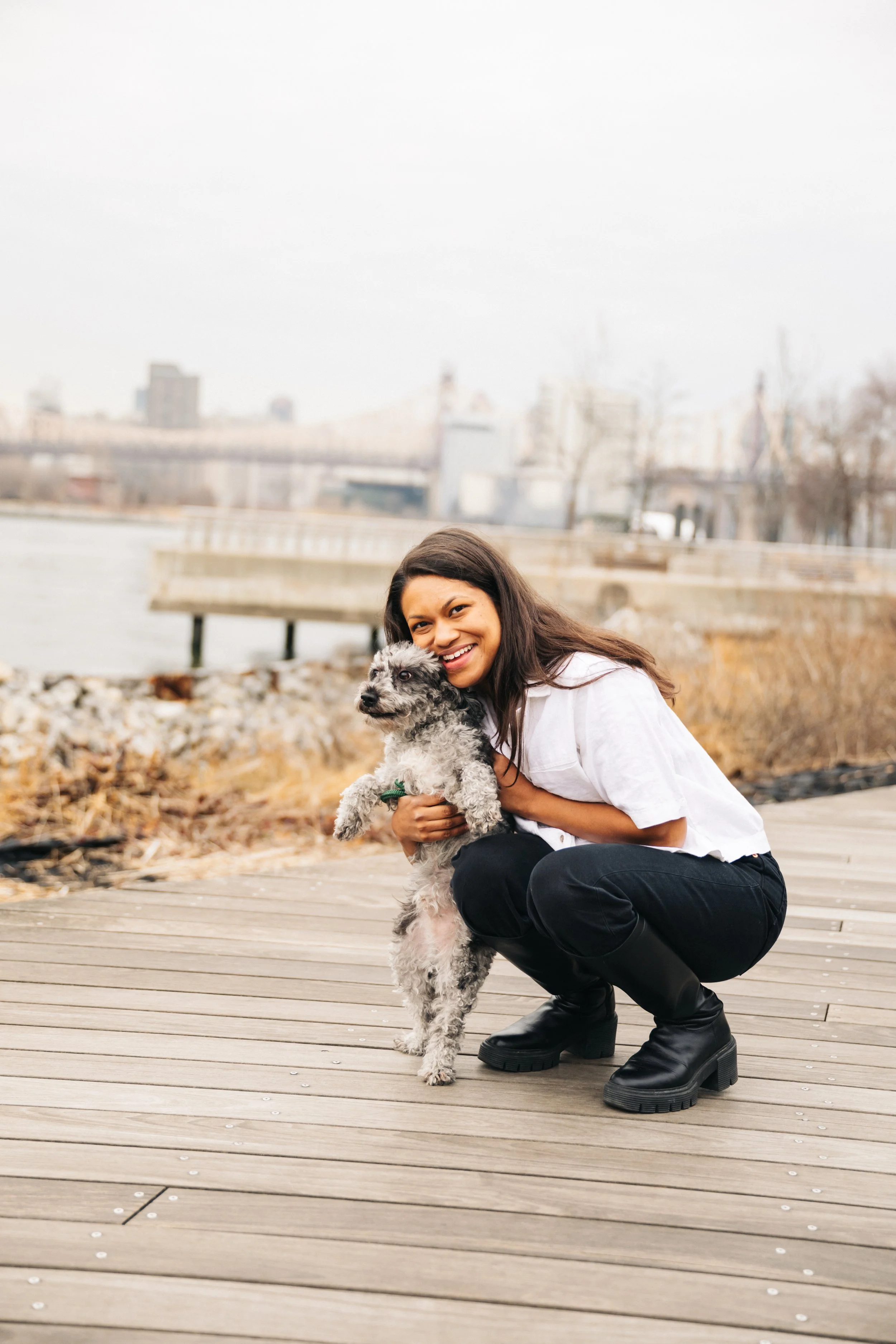 A woman crouching on a wooden dock while holding a gray and white dog near a body of water with an urban skyline in the background.